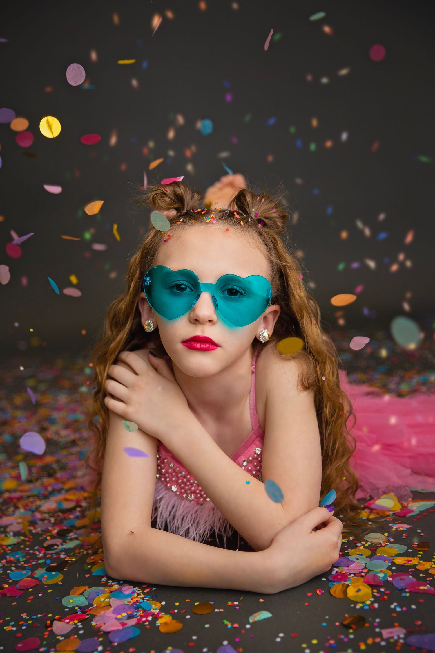 Girl in pink dress and teal heart sunglasses lying among colorful confetti.