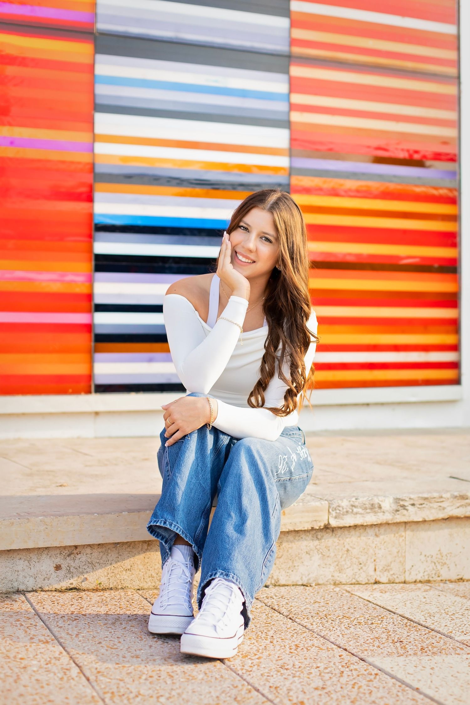 Teenage girl in white off-shoulder top and jeans sits smiling before colorful striped art installation.