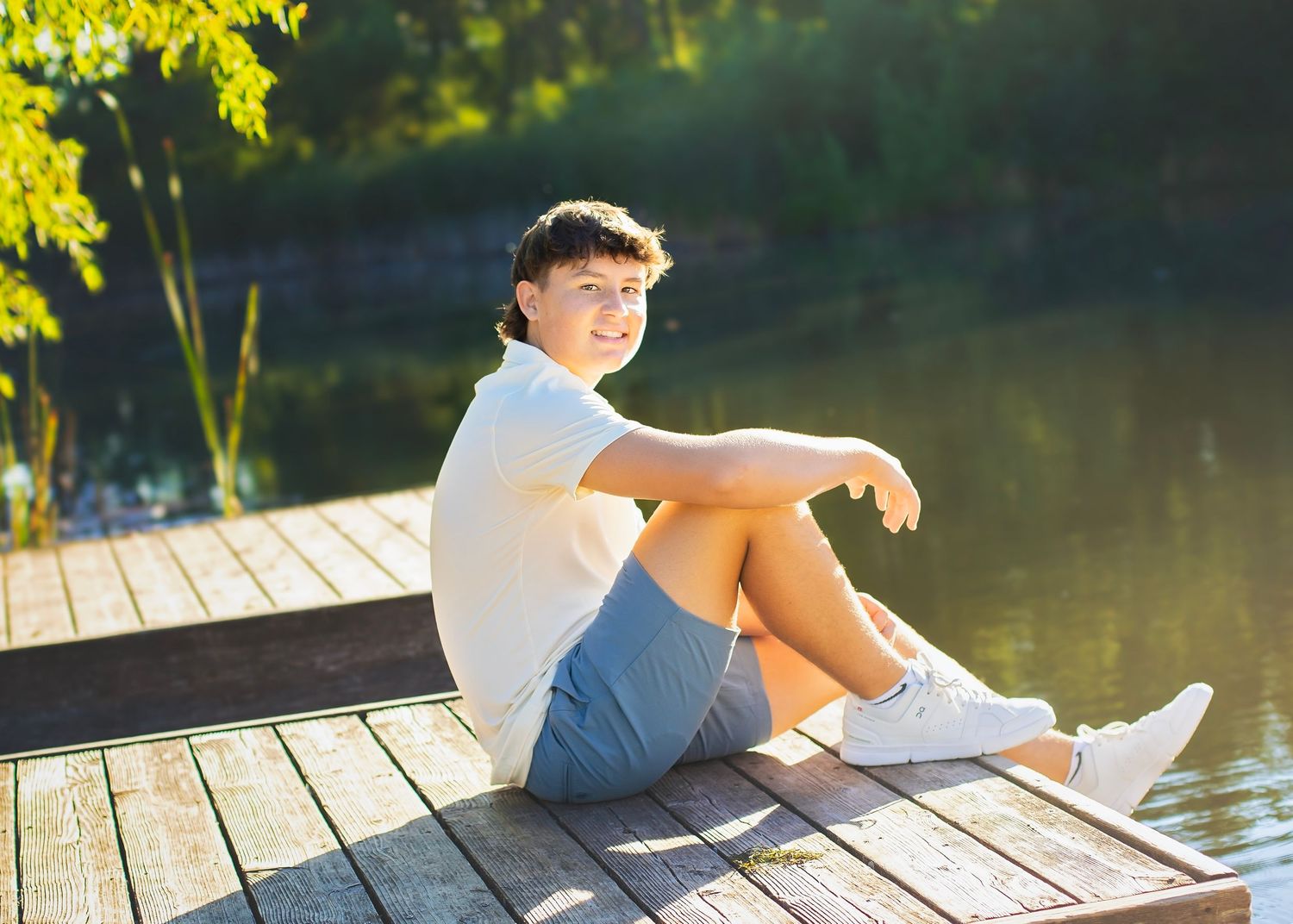 Happy teenage boy sitting on a sunny wooden dock by a calm reflective lake smiling at camera.