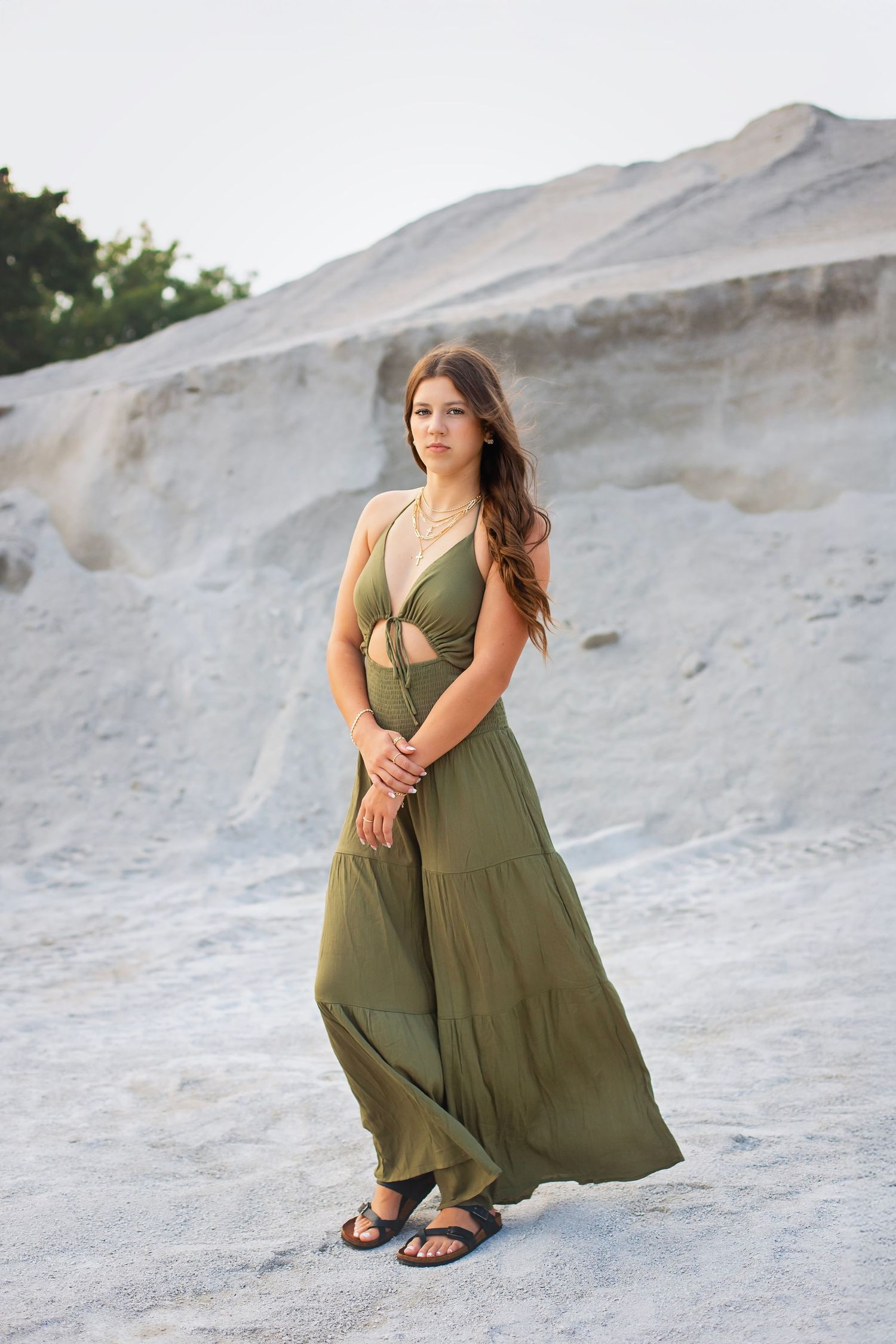Young woman in flowing olive green halter maxi dress stands gracefully in a sandy quarry landscape.
