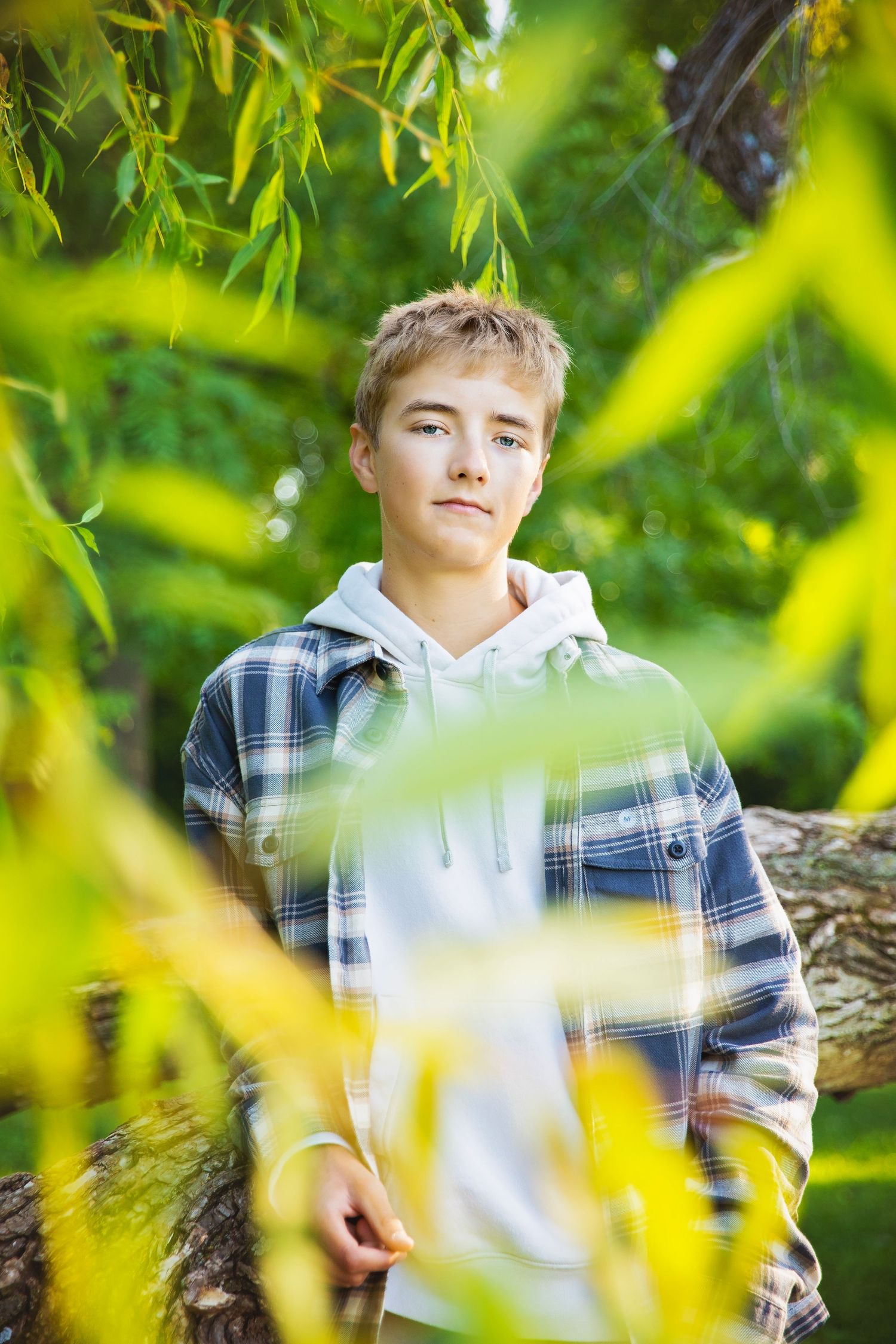Teen boy in plaid shirt and hoodie leaning against tree trunk surrounded by green foliage outdoors.
