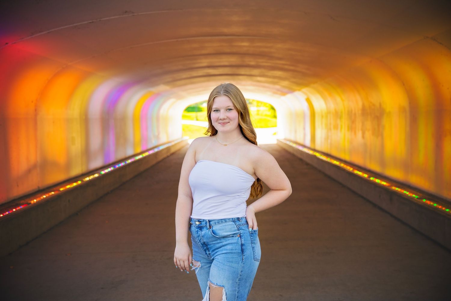 Smiling teen girl in white top and jeans poses inside a colorful illuminated tunnel.