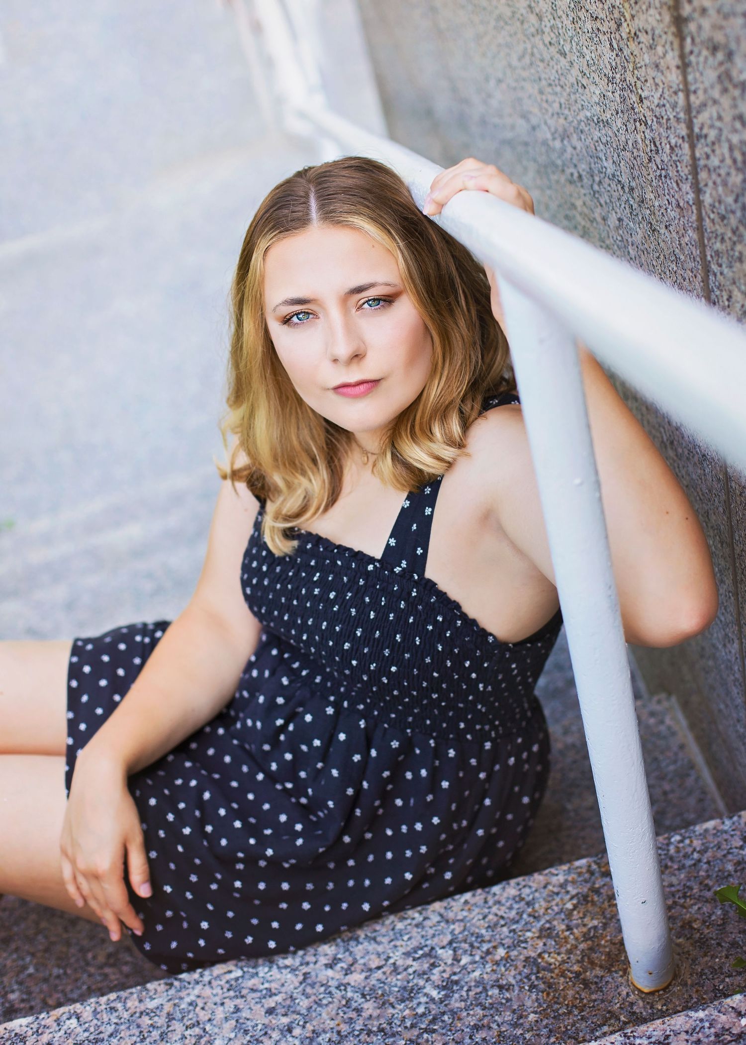 Blonde teen girl in navy polka dot dress sitting on outdoor stone steps holding white railing.
