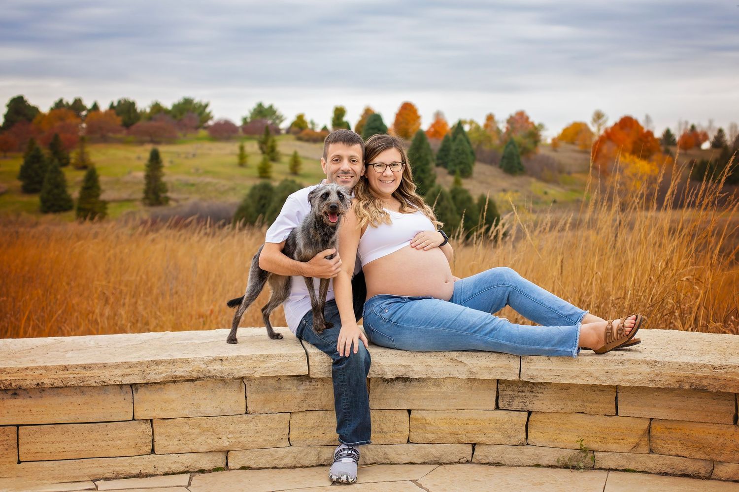 Pregnant woman in jeans and man with gray dog sit together on stone wall in autumn field.