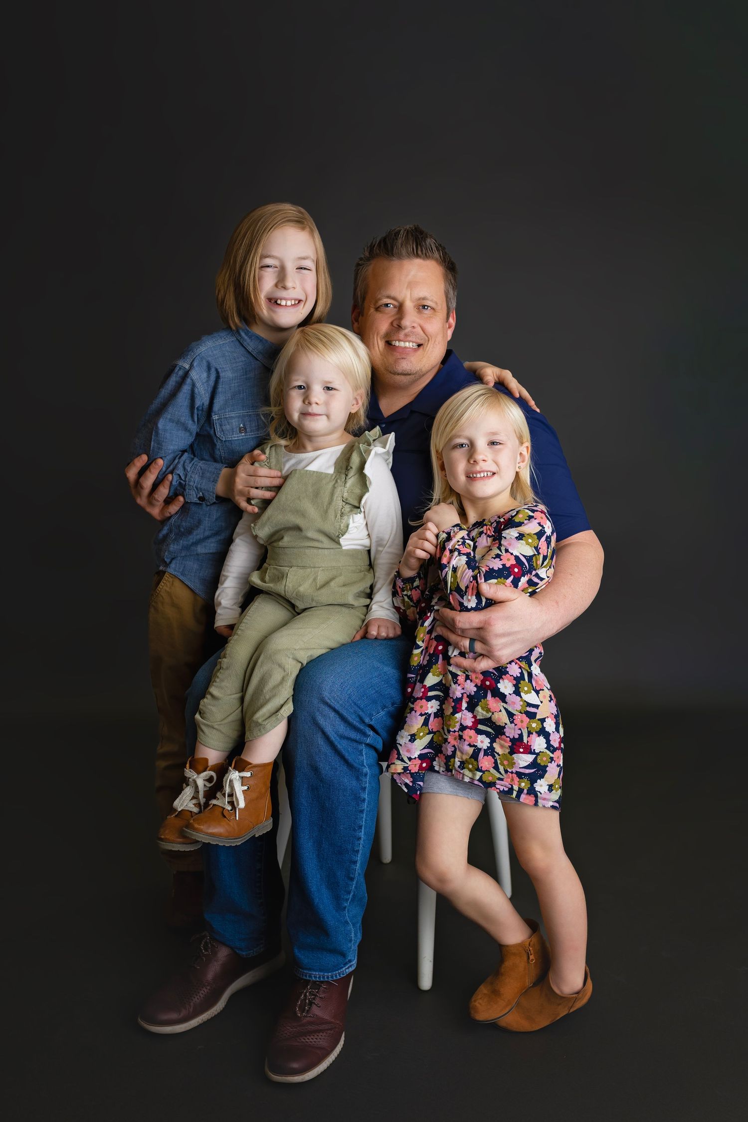 Cheerful father sitting on white chair surrounded by three happy children in dark background studio photo.