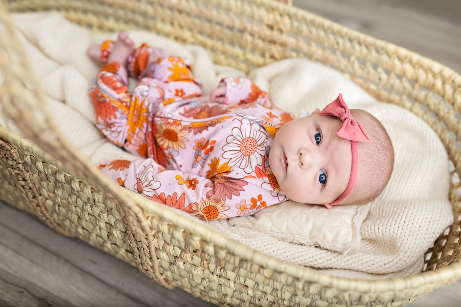 Newborn baby girl in floral pink outfit with bow headband lying in woven Moses basket.