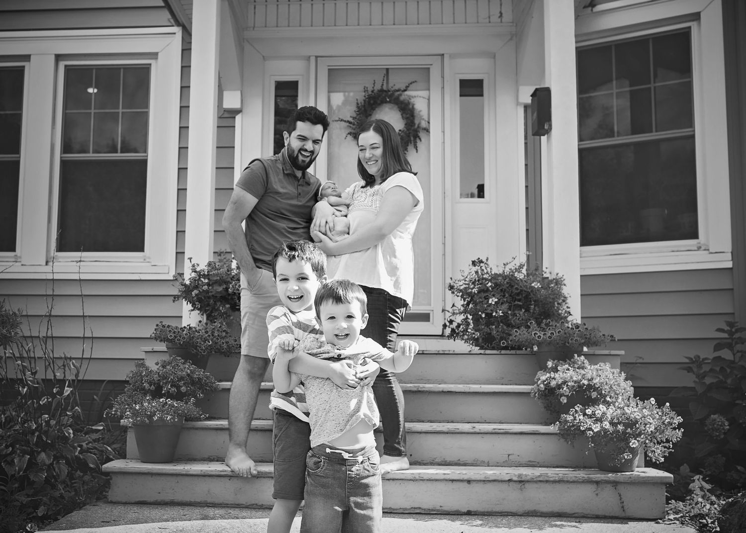 Black and white family photo of parents with toddler and newborn standing on home front steps.