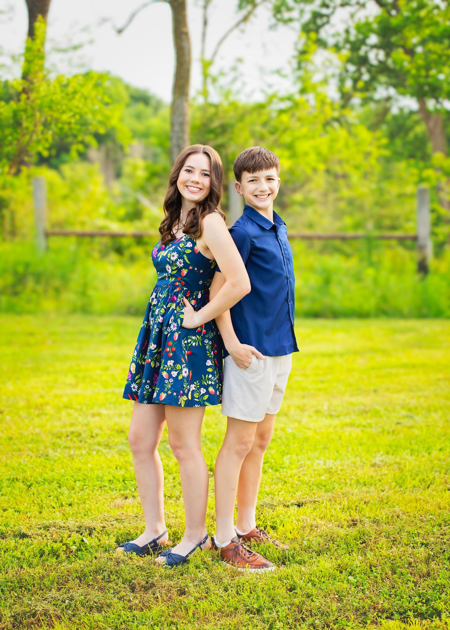 Smiling teenage girl and boy pose back-to-back in a sunny green outdoor setting.