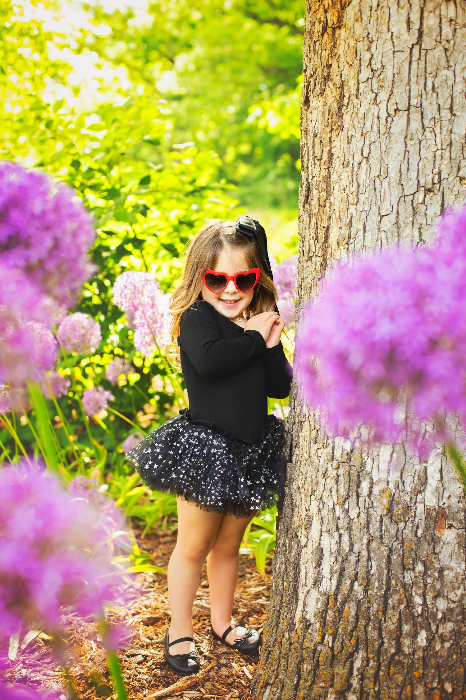 Adorable girl in black tutu and heart sunglasses standing by tree surrounded by purple flowers.