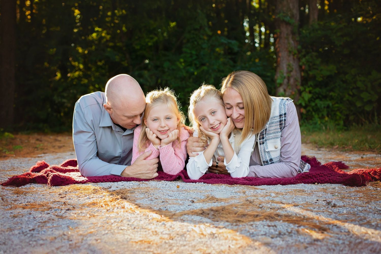 Family of four lying on a blanket outdoors, smiling, with two young girls between their parents in a forest setting.