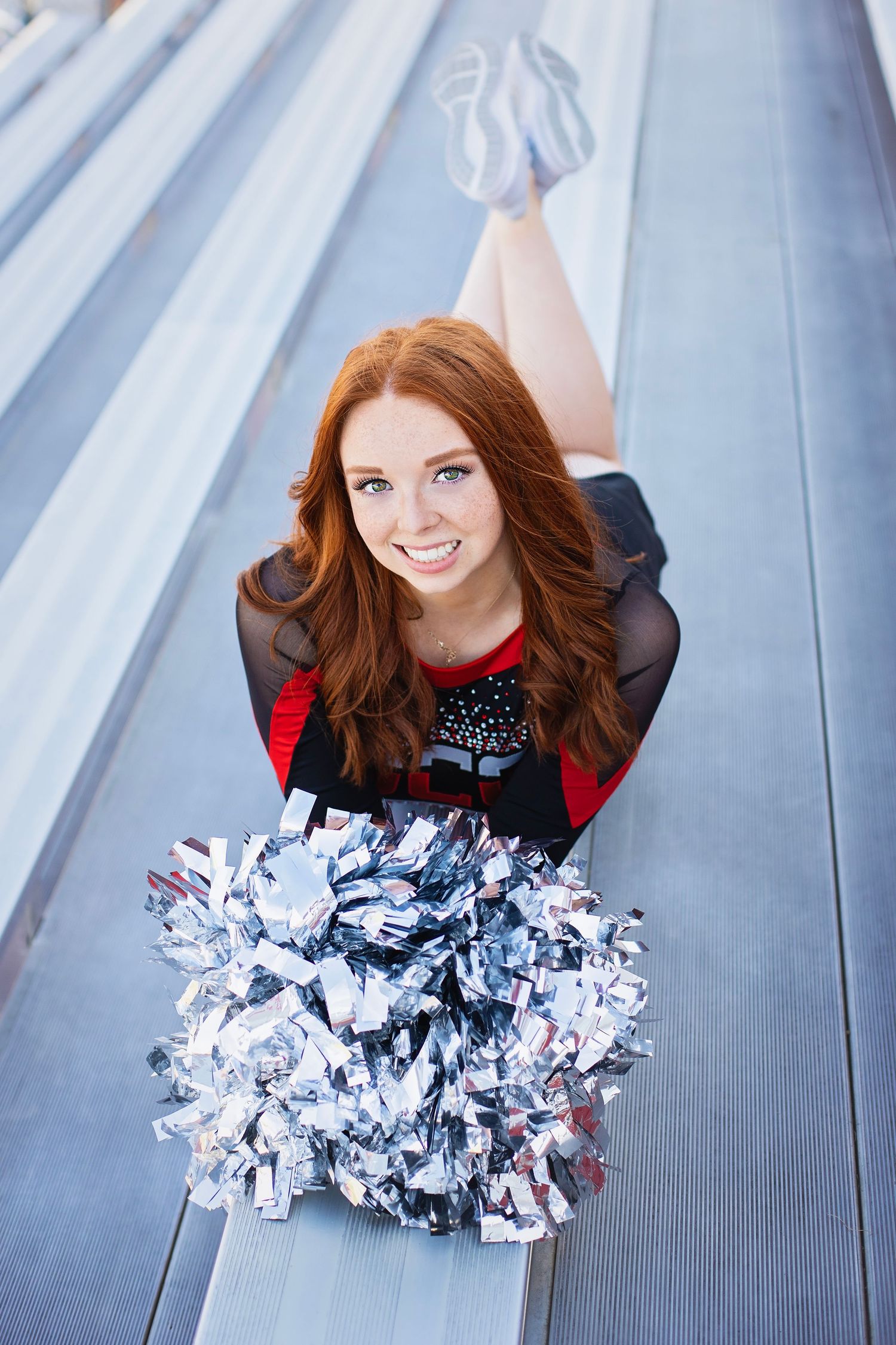 Cheerful redhead cheerleader in uniform holds silver pom-poms while posing on bleachers with leg raised.
