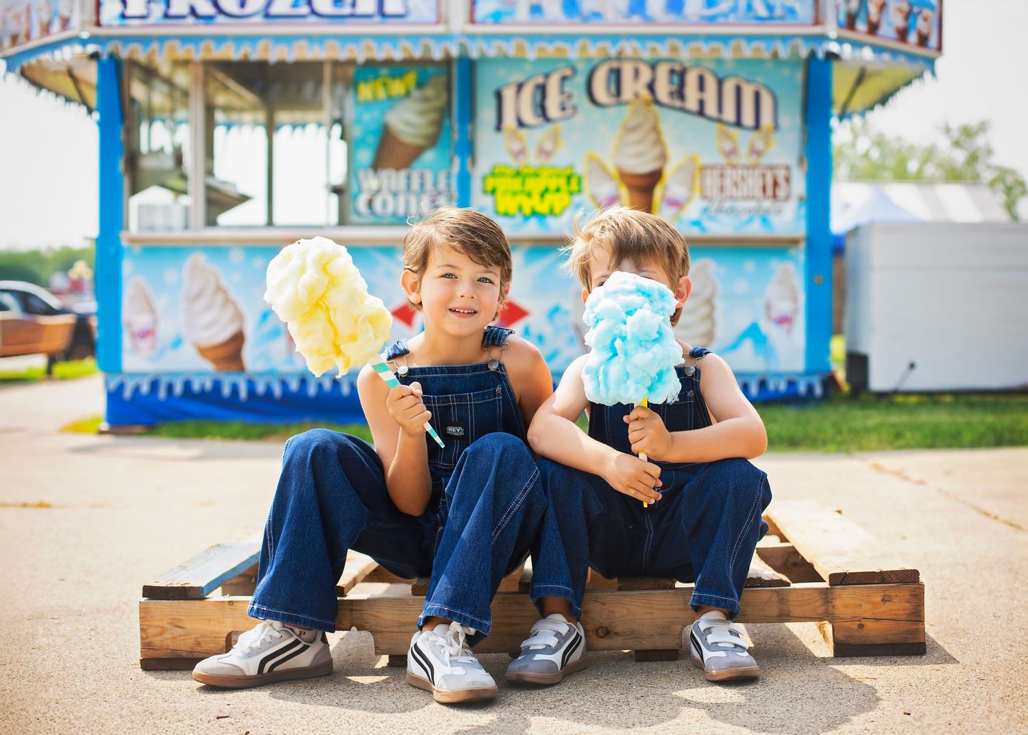 Two young boys in denim overalls eating cotton candy in front of a fair ice cream stand.