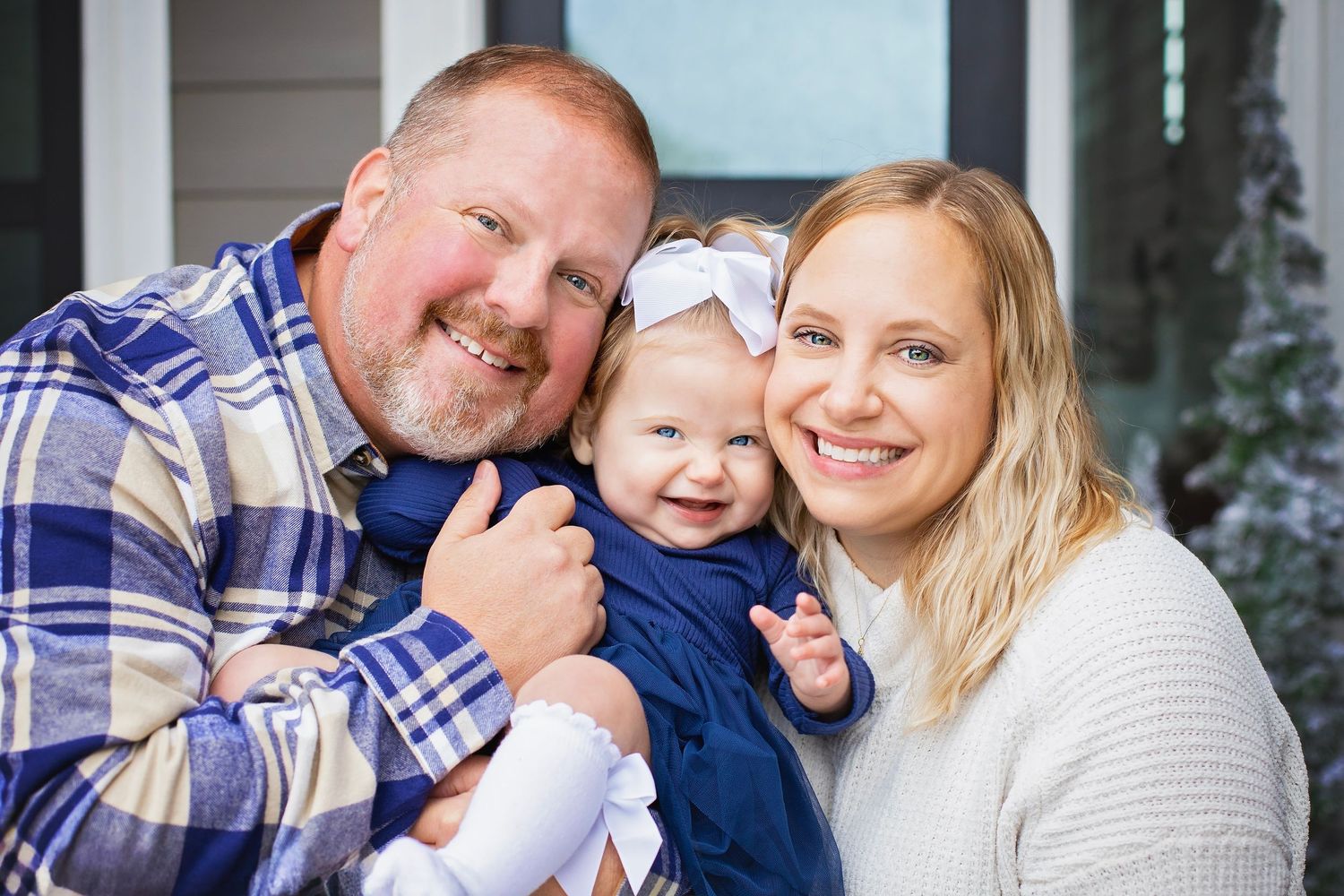 Loving family portrait with parents and smiling baby girl in a navy dress with white bow.