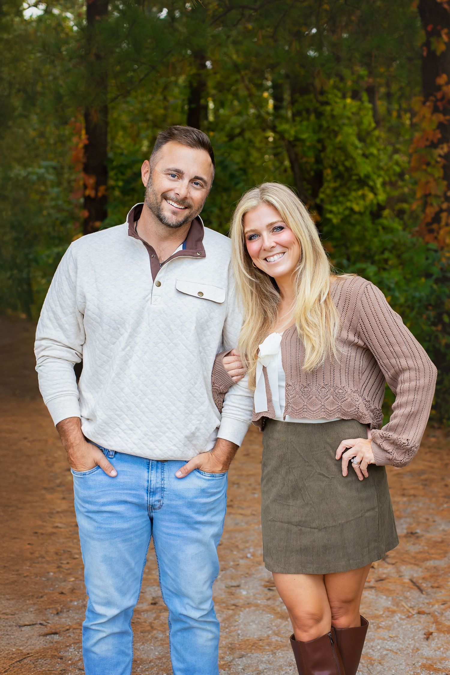 A smiling couple stands together on a forest path surrounded by colorful autumn foliage.
