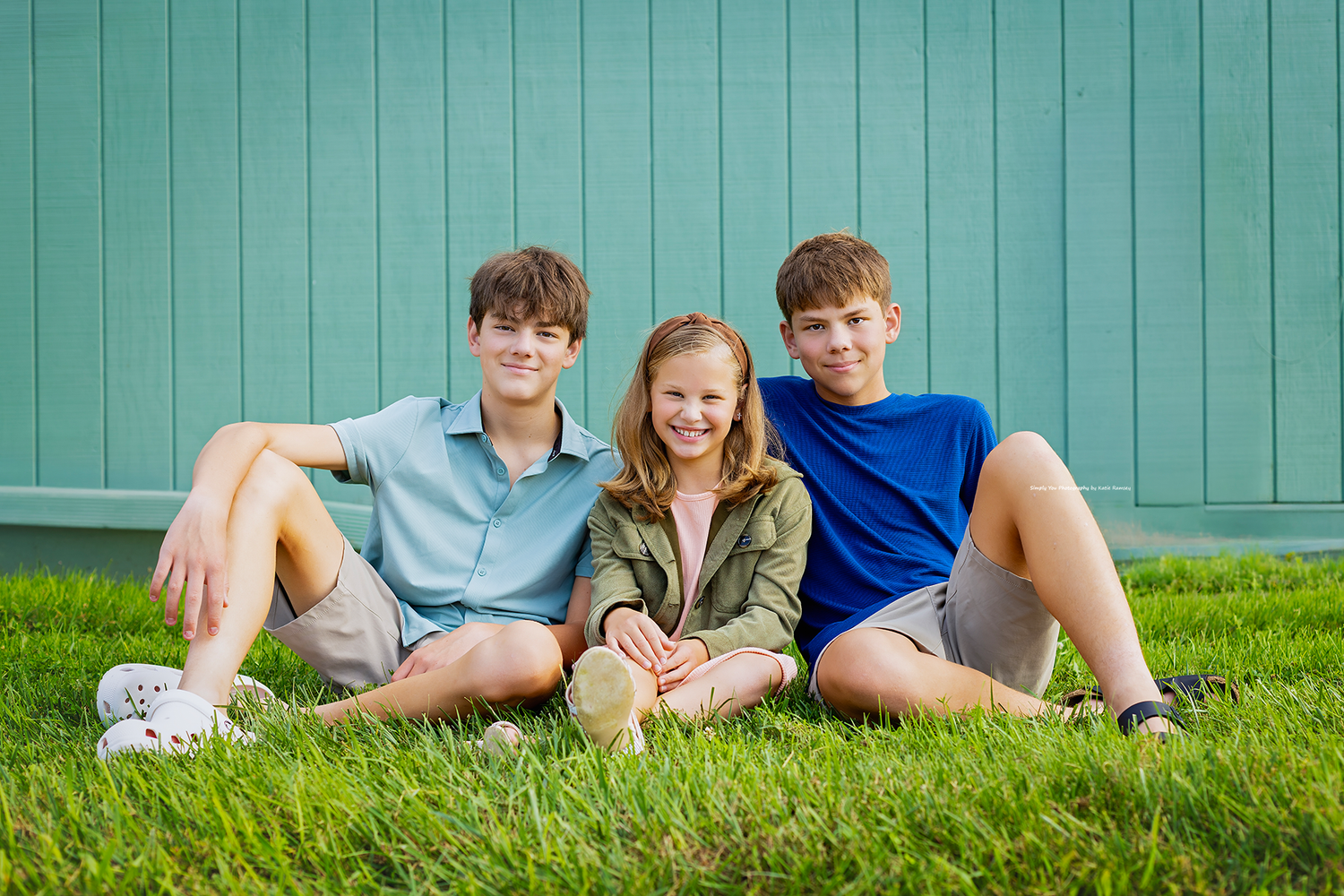 Three smiling children sitting together on green grass in front of a teal wall outdoors.