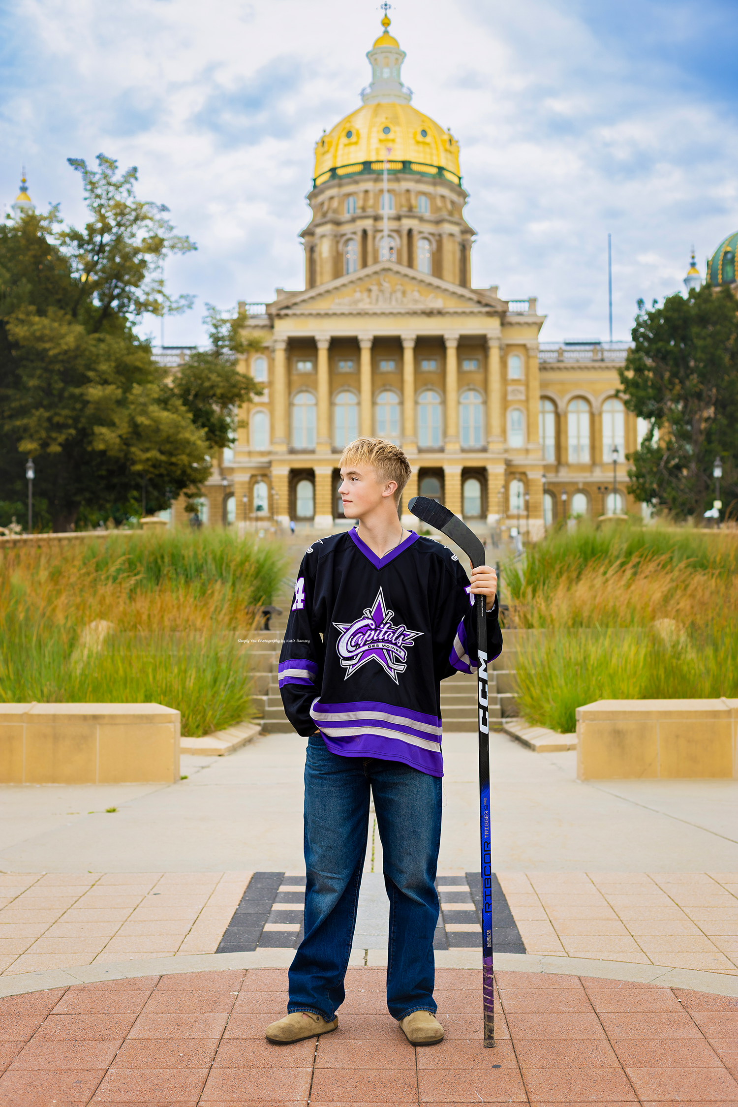Young hockey player in purple jersey holds stick before Iowa State Capitol building with golden dome.