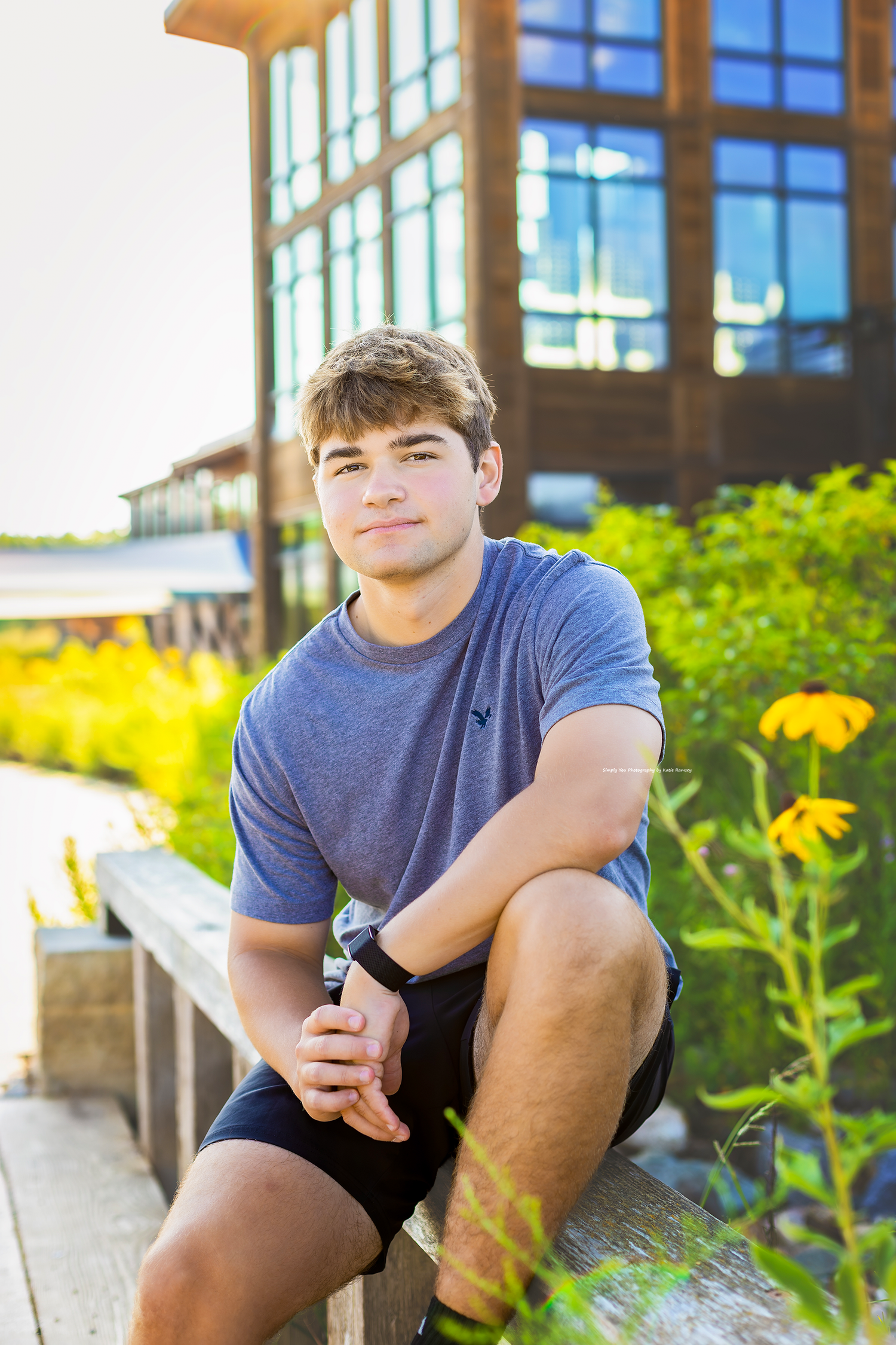 Teen boy in blue t-shirt sitting outdoors near yellow flowers by a modern building.