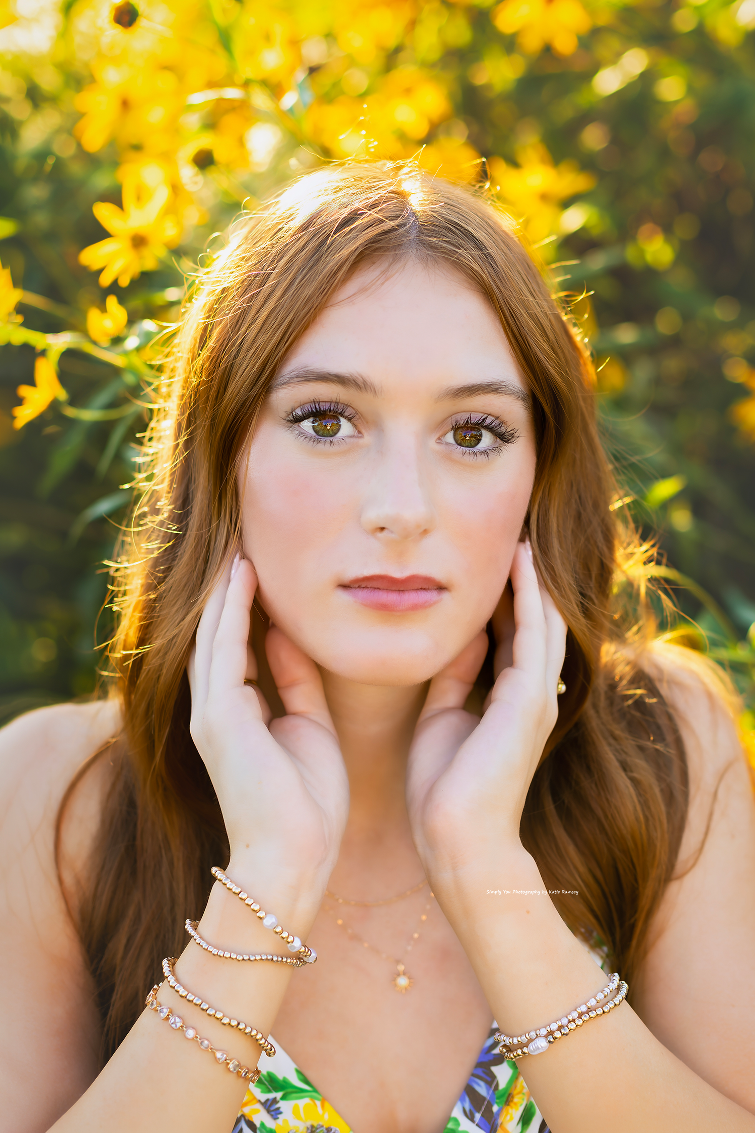 Young woman with red hair and bracelets poses with hands on face near vibrant yellow flowers outdoors.