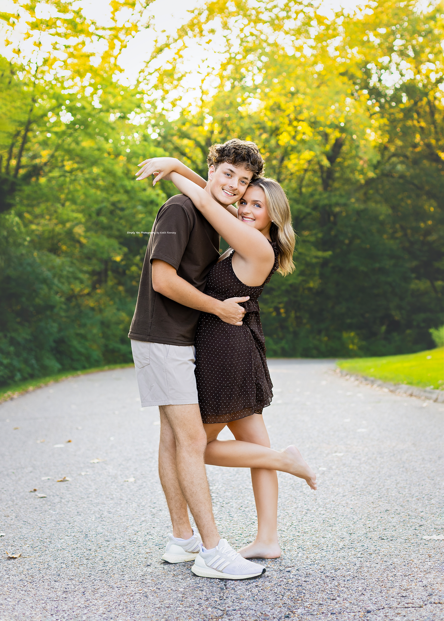 A young couple hugging and smiling on a tree-lined path, wearing casual summer outfits in a lush green park.
