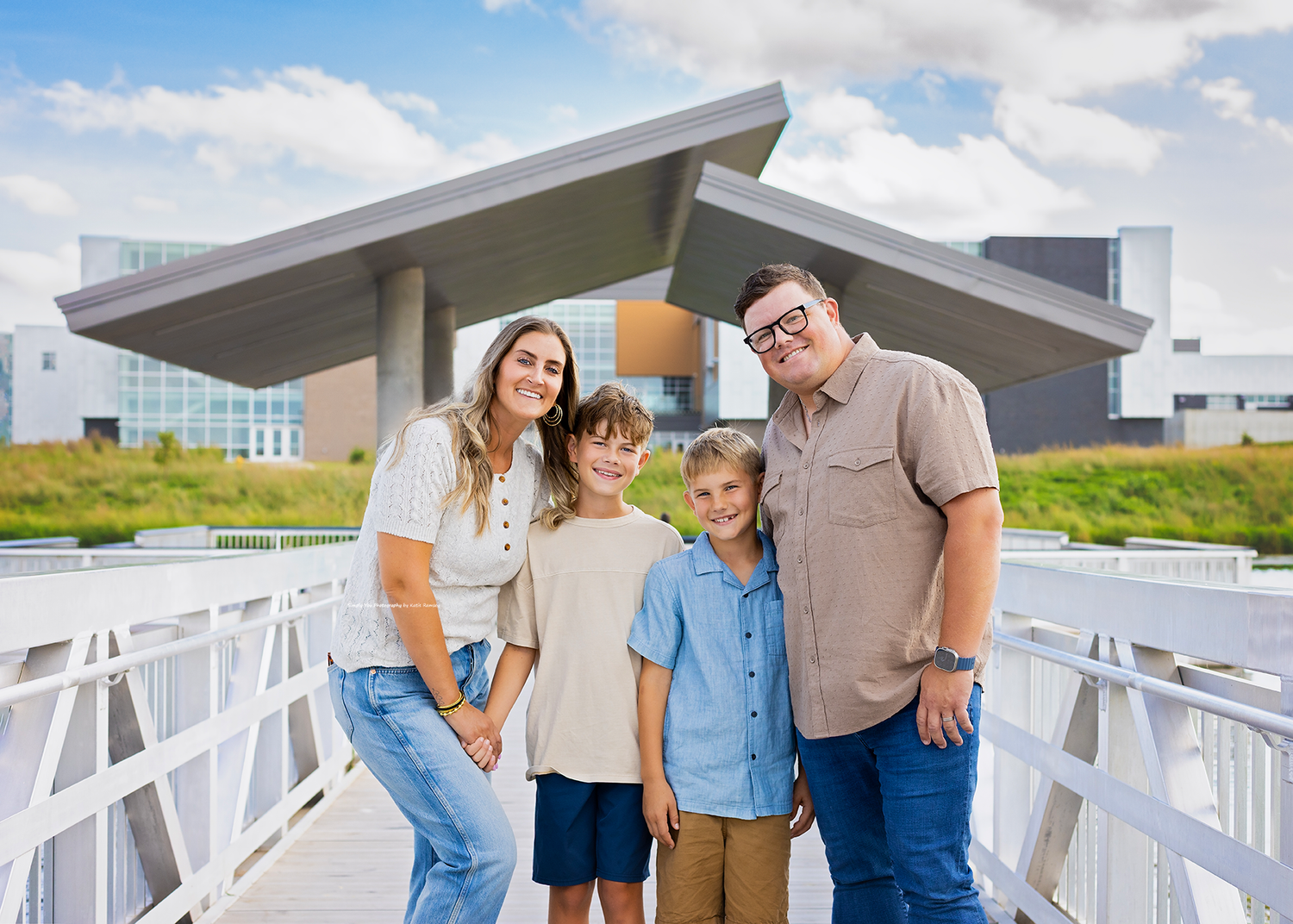 Family of four smiling together on a bridge outside a modern building on a sunny day.