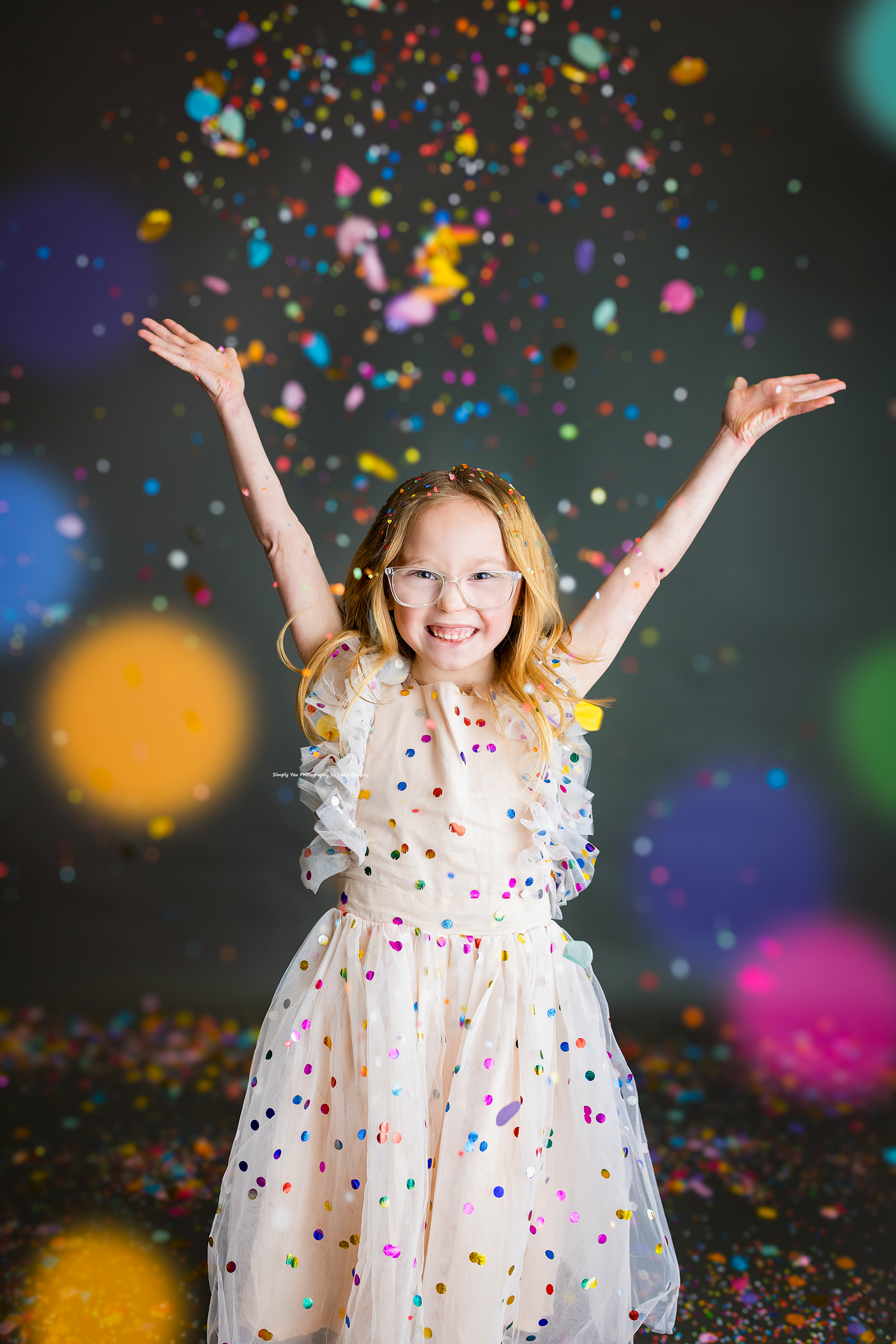 Excited young girl in a polka dot dress throwing colorful confetti with arms raised in a studio.