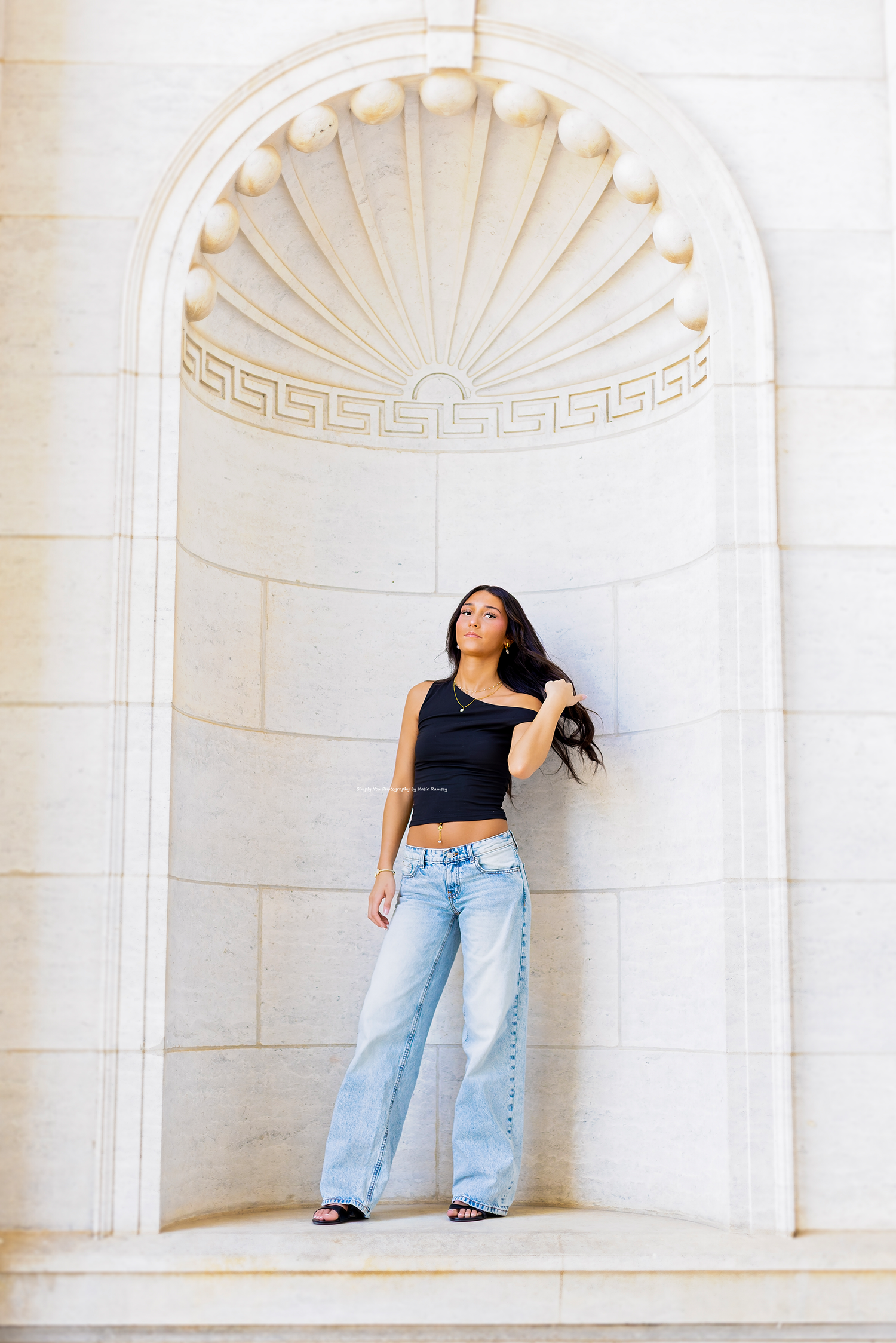 Young woman in black crop top and wide-leg jeans posing in ornate white stone archway.