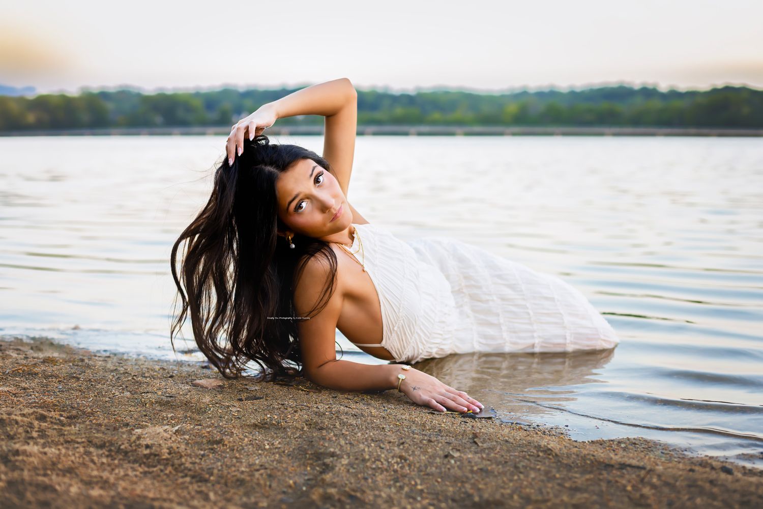A young woman in a white dress poses gracefully by a calm lake shore at dusk with her hair flowing.