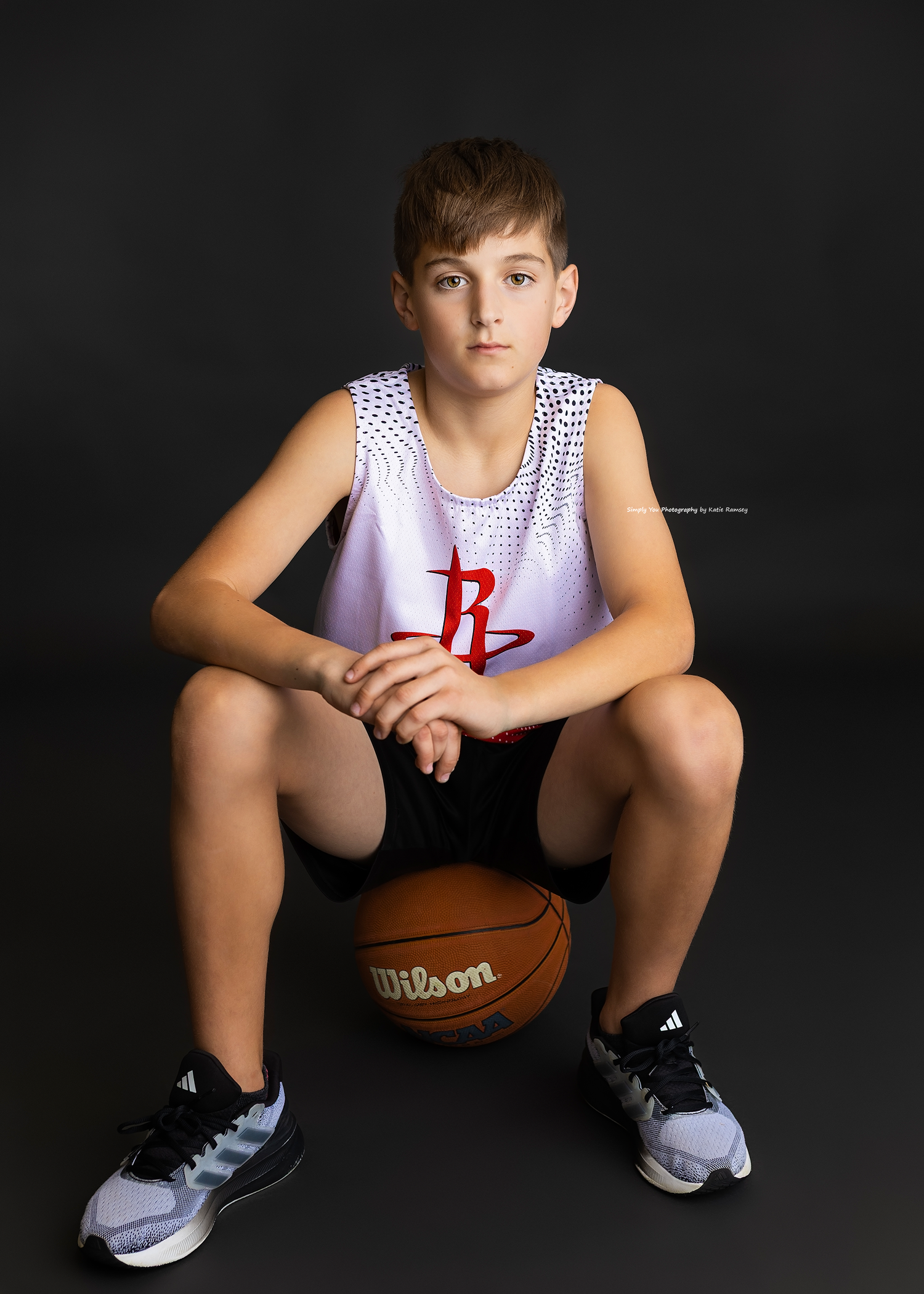 Young boy in Houston Rockets jersey sits on Wilson basketball for sports portrait photo.