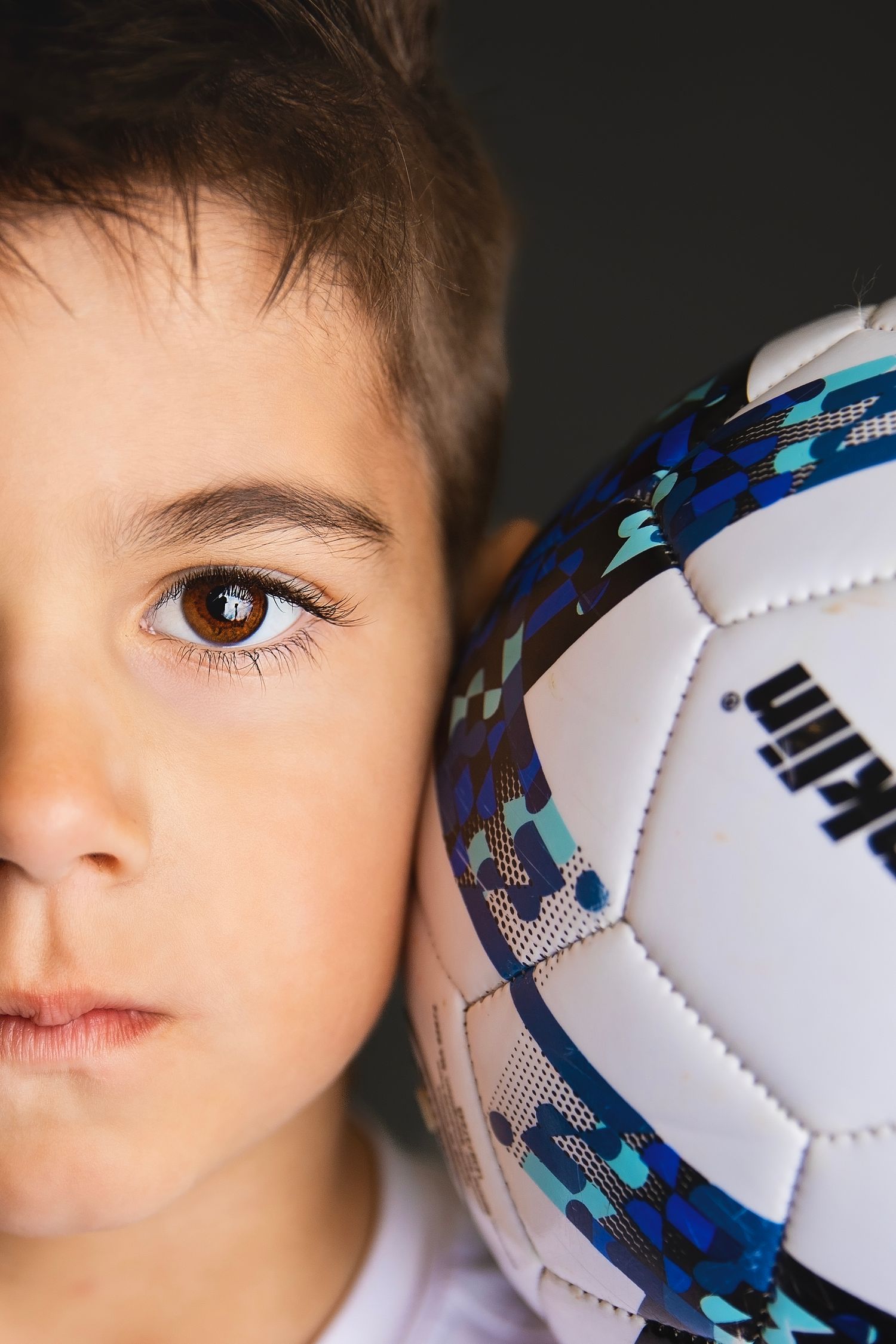 Close-up portrait of young boy with brown eyes holding a Kipsta soccer ball beside his face.
