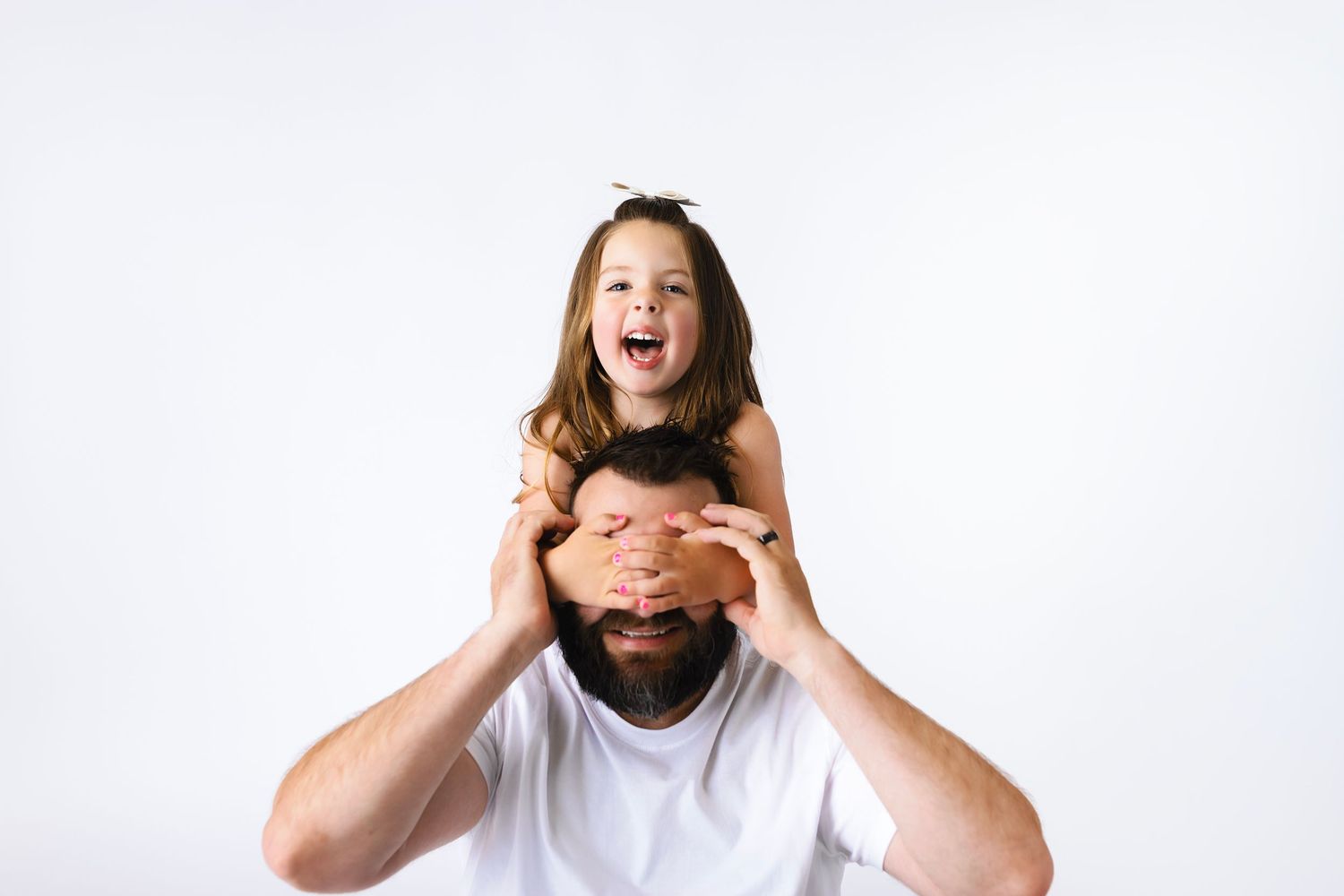 A laughing girl sits on her father's shoulders covering his eyes against a white background.