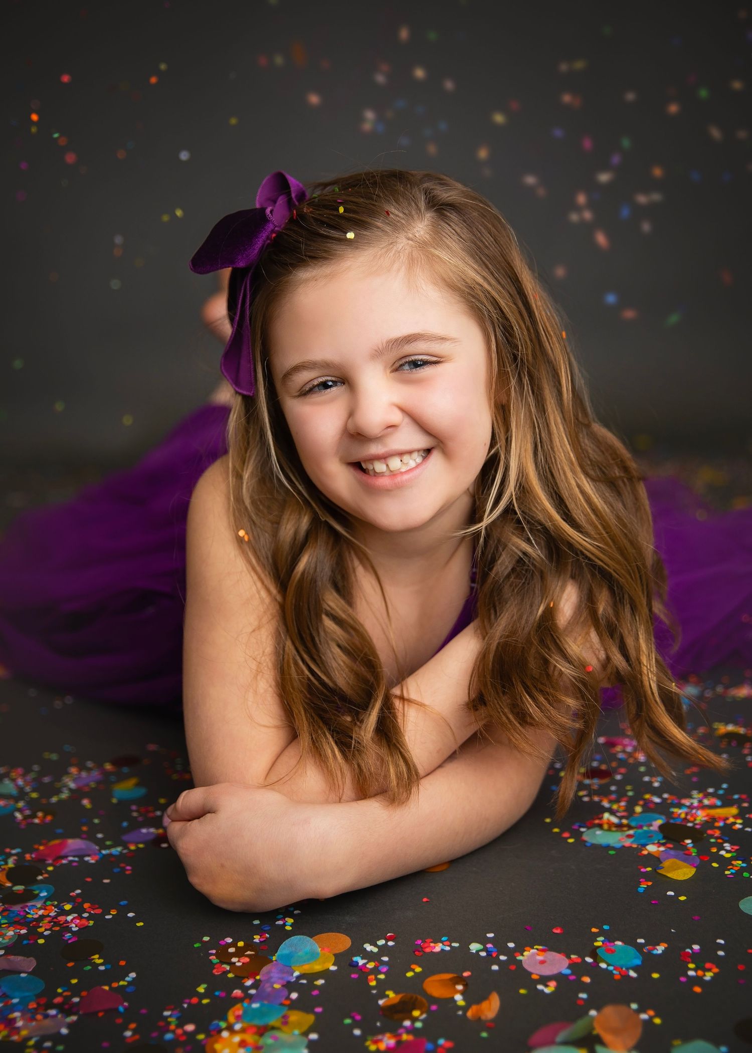 Cheerful girl in purple tutu lying down smiling, surrounded by colorful confetti on dark background.