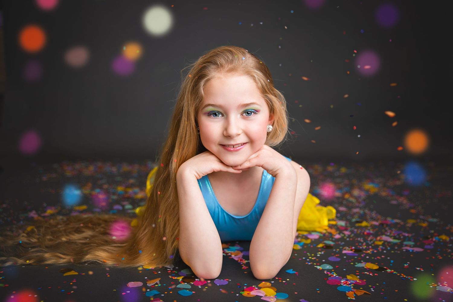 A cheerful young girl in a blue top rests her chin on her hands while lying among colorful confetti on a dark floor.