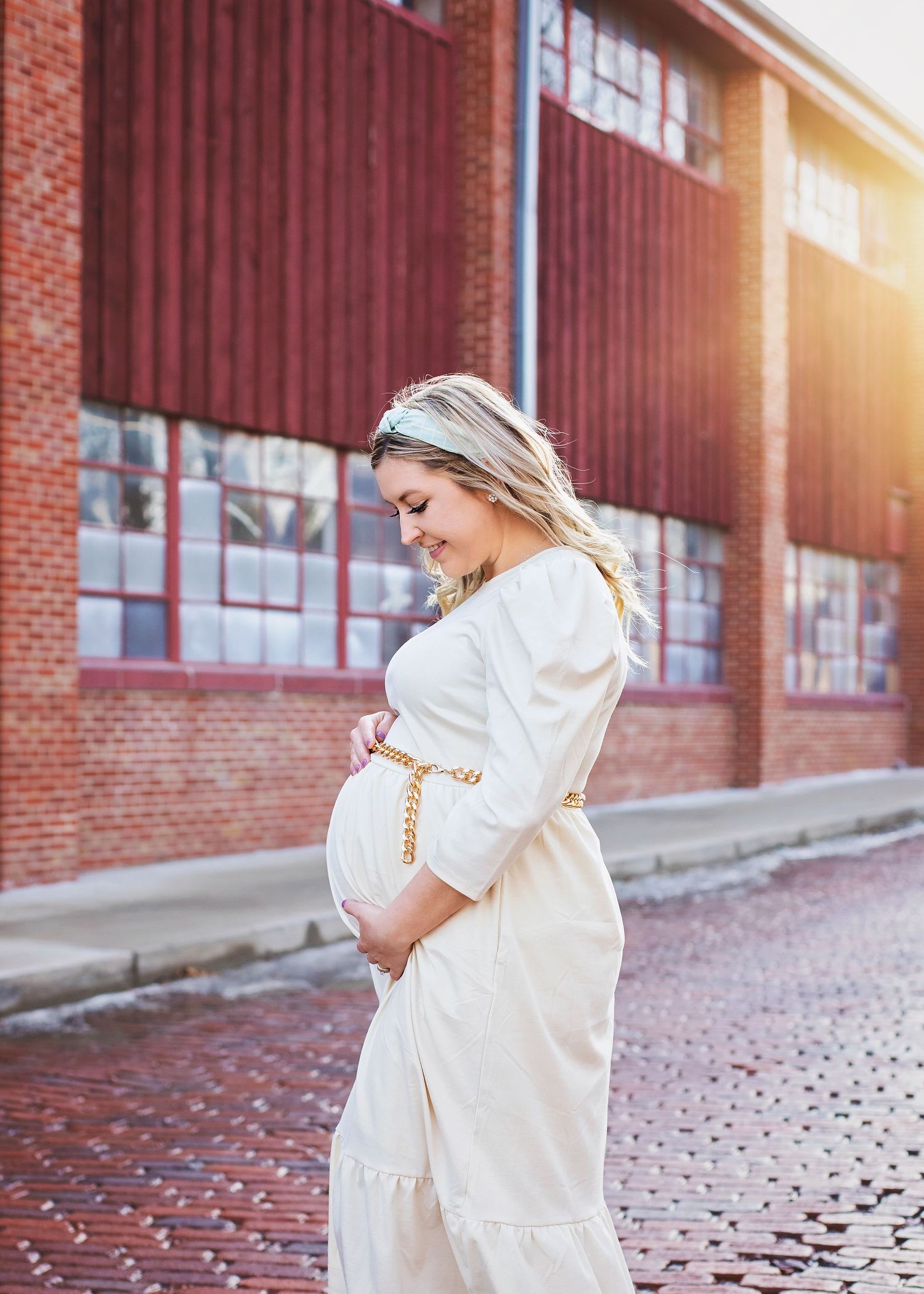 Pregnant woman in white dress with gold belt cradling baby bump outdoors.