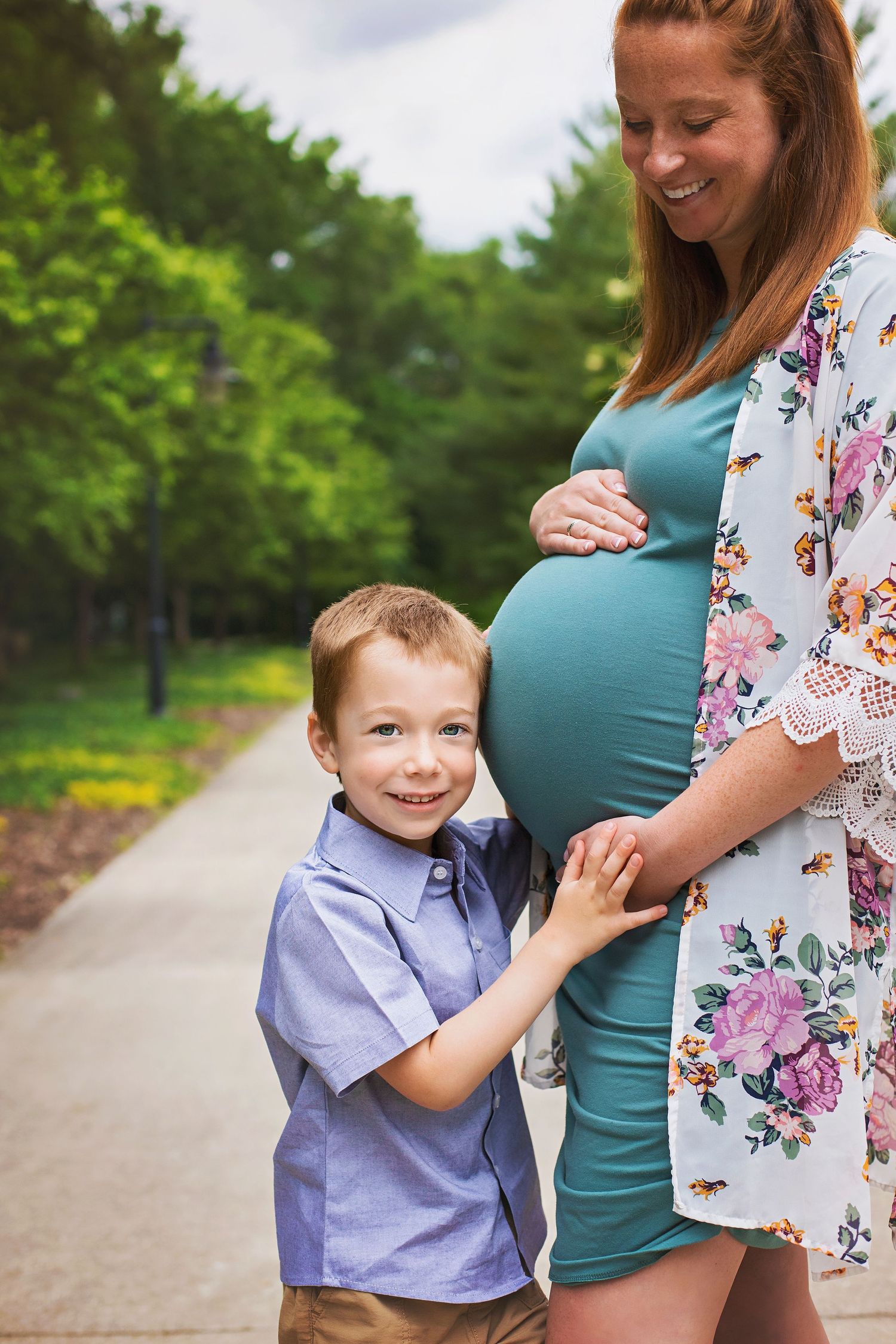 Young boy hugs his pregnant mothers baby bump outdoors on a sunny day, both smiling joyfully.