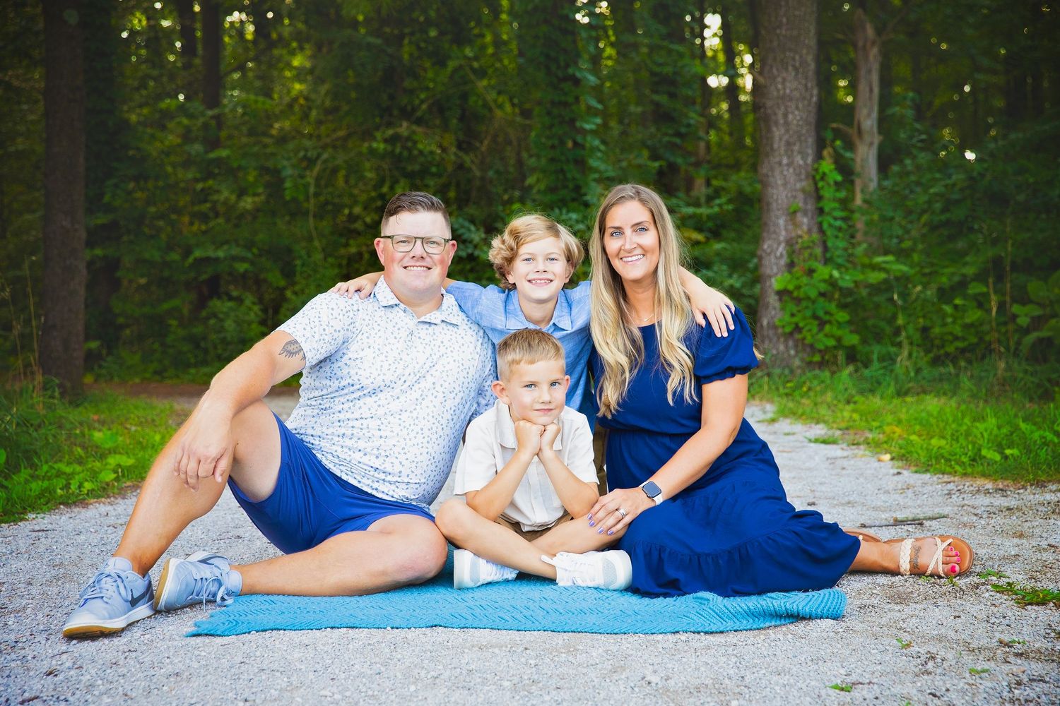 A family of four sits casually on a blanket along a wooded path in warm summer light.