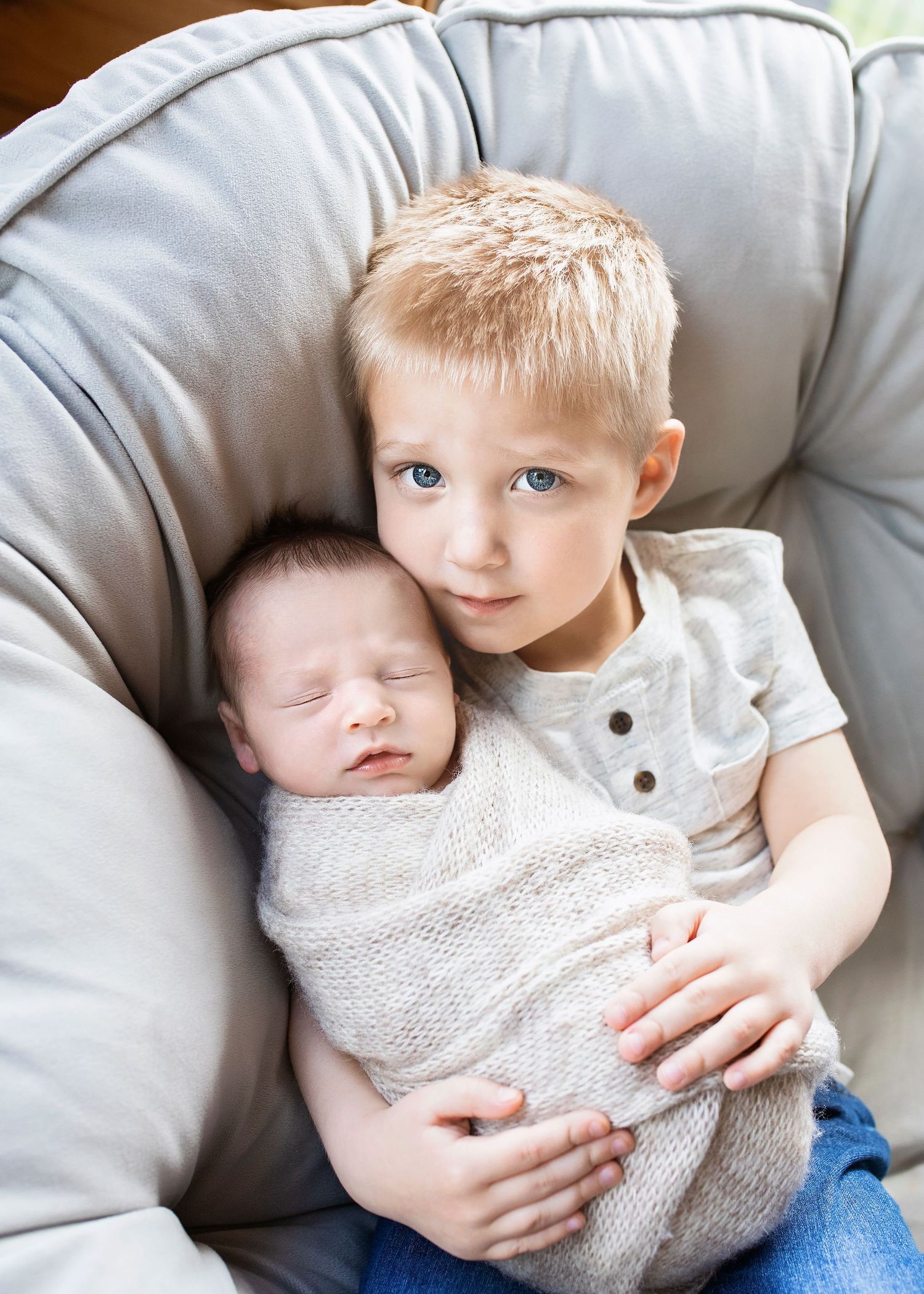 Young blonde boy gently holding sleeping swaddled newborn sibling in a cozy gray chair.