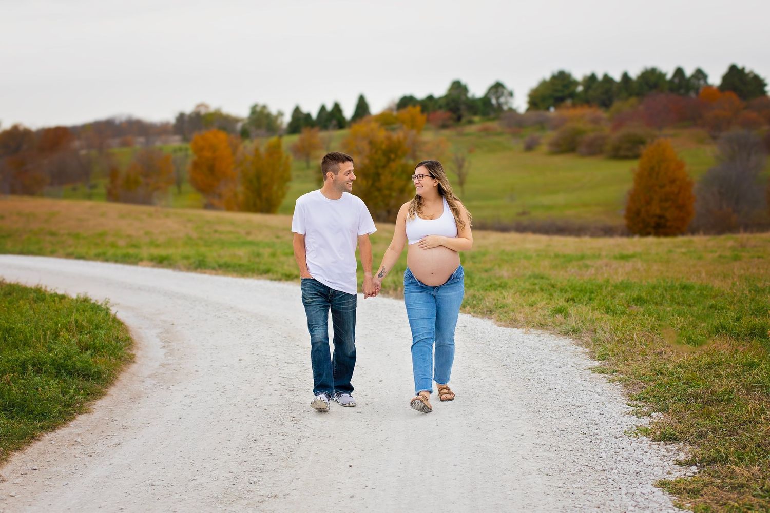 Expecting couple walks hand-in-hand down a winding gravel path surrounded by autumn foliage.