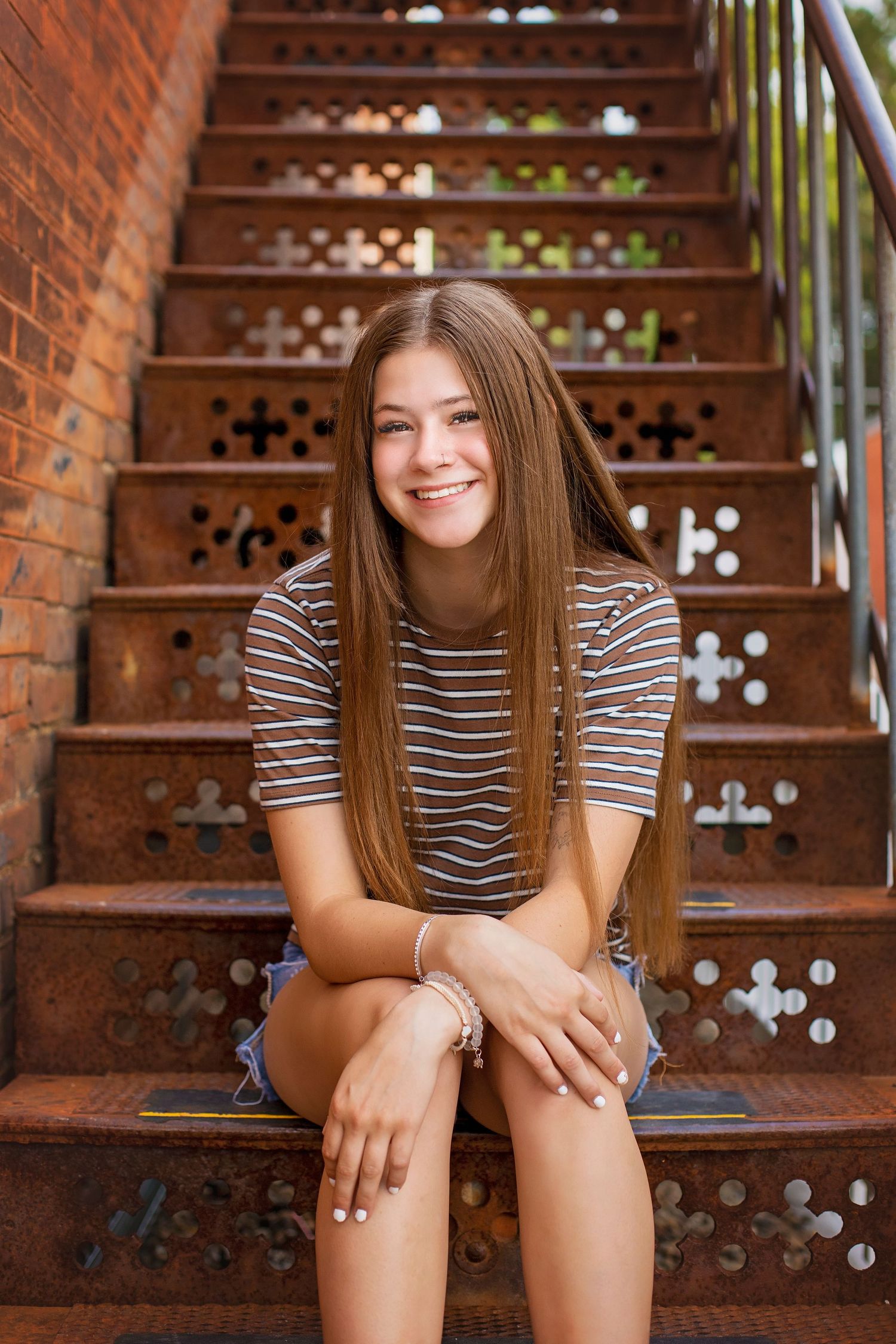 Smiling teen girl with long brown hair sitting on rustic metal stairs in striped shirt and denim shorts.