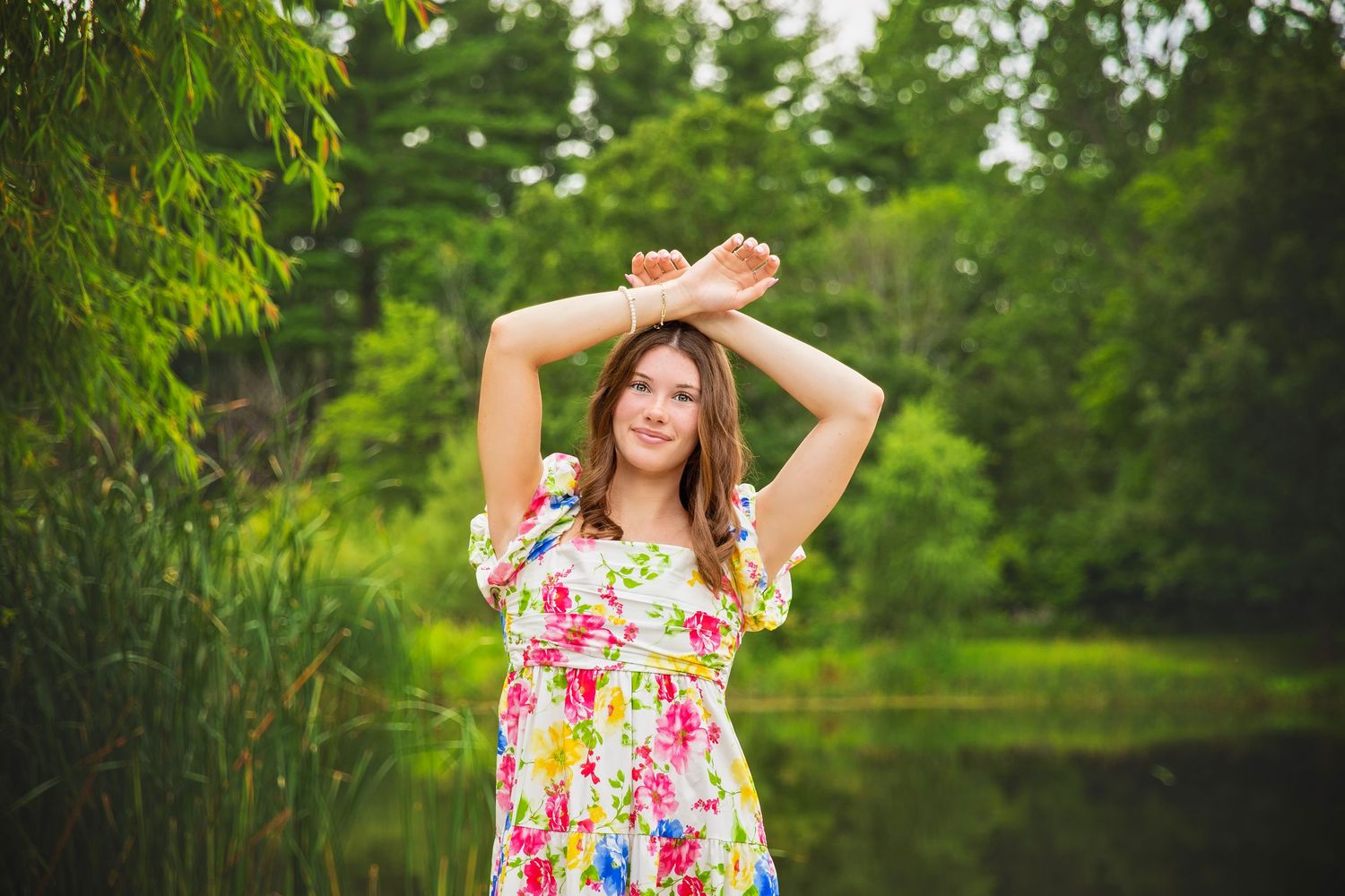 Teen girl in colorful floral dress posing with arms raised near a lush green pond.
