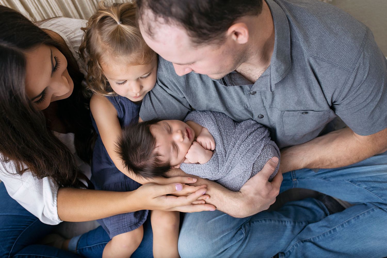 Family of four with parents and young daughter admiring their newborn baby wrapped in gray swaddle.