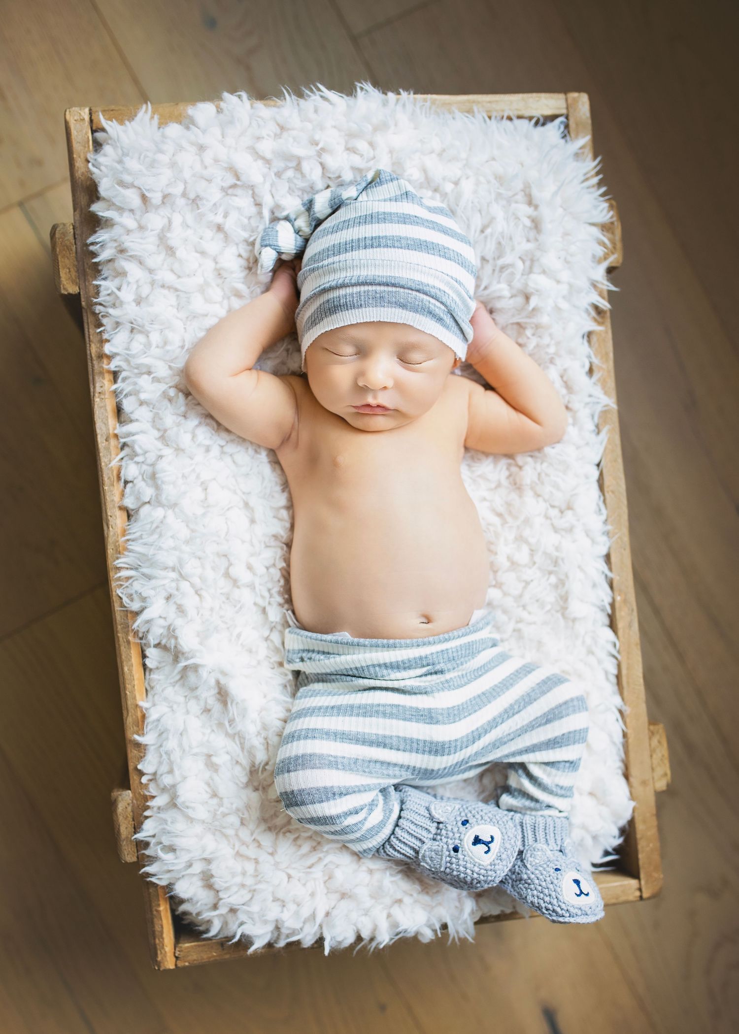 Sleeping newborn baby boy in striped pants and knotted hat lying on fluffy white rug in wooden crate.