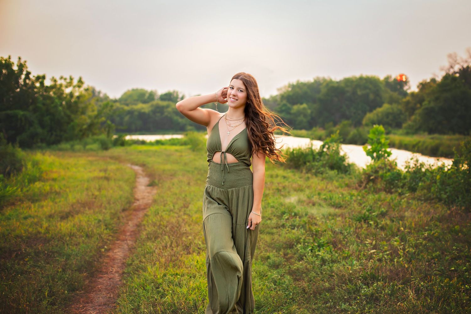 Young woman in olive green jumpsuit walks along scenic nature path at sunset.