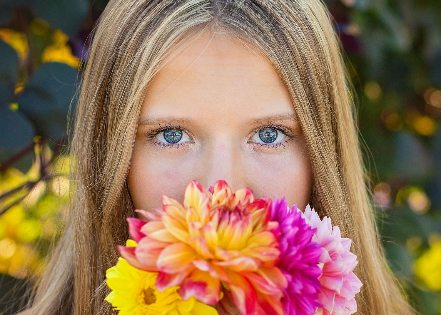 Close-up portrait of blue-eyed blonde girl holding colorful dahlia bouquet over her face outdoors.
