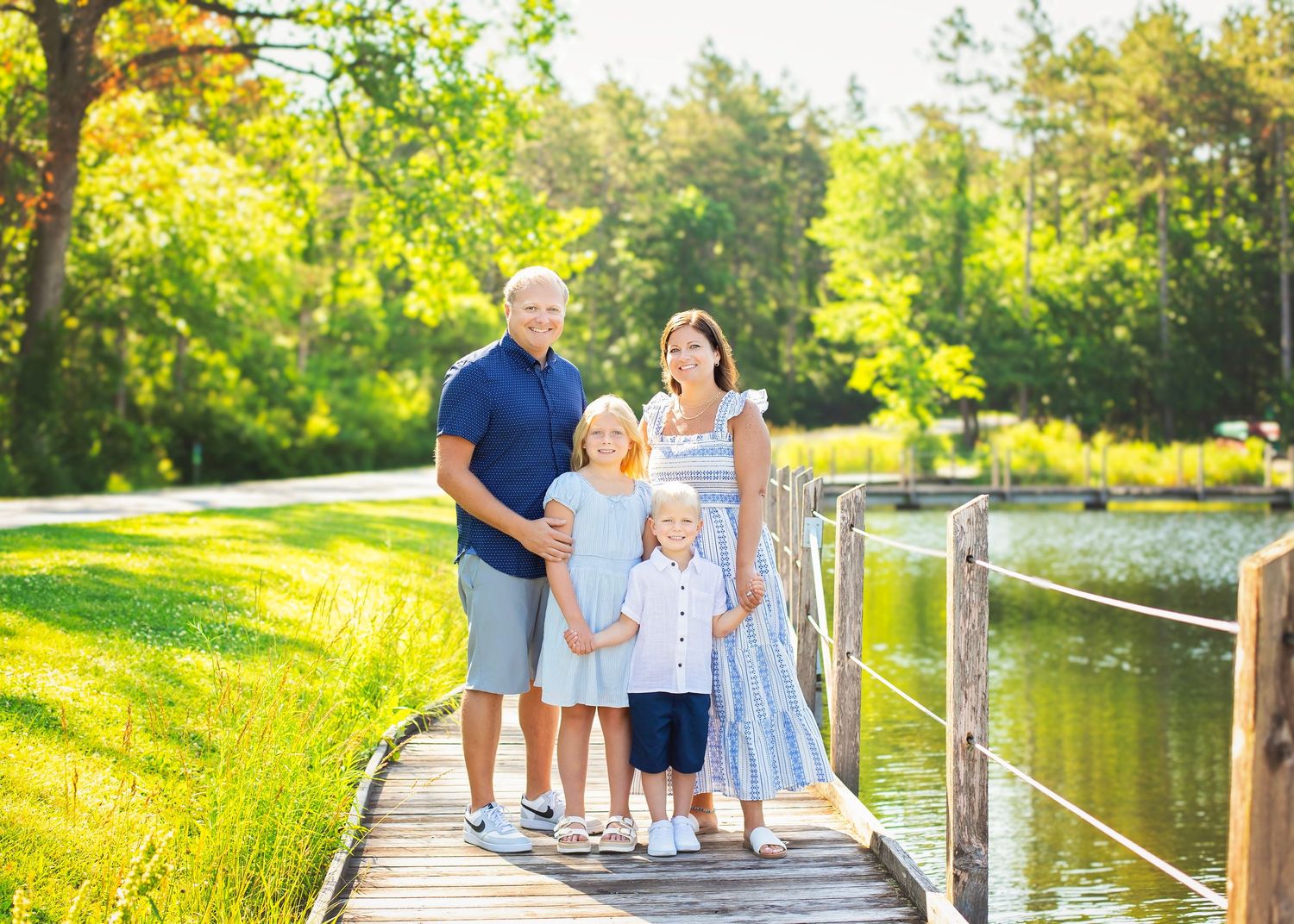 Family of four standing on wooden dock beside calm water surrounded by lush green trees.