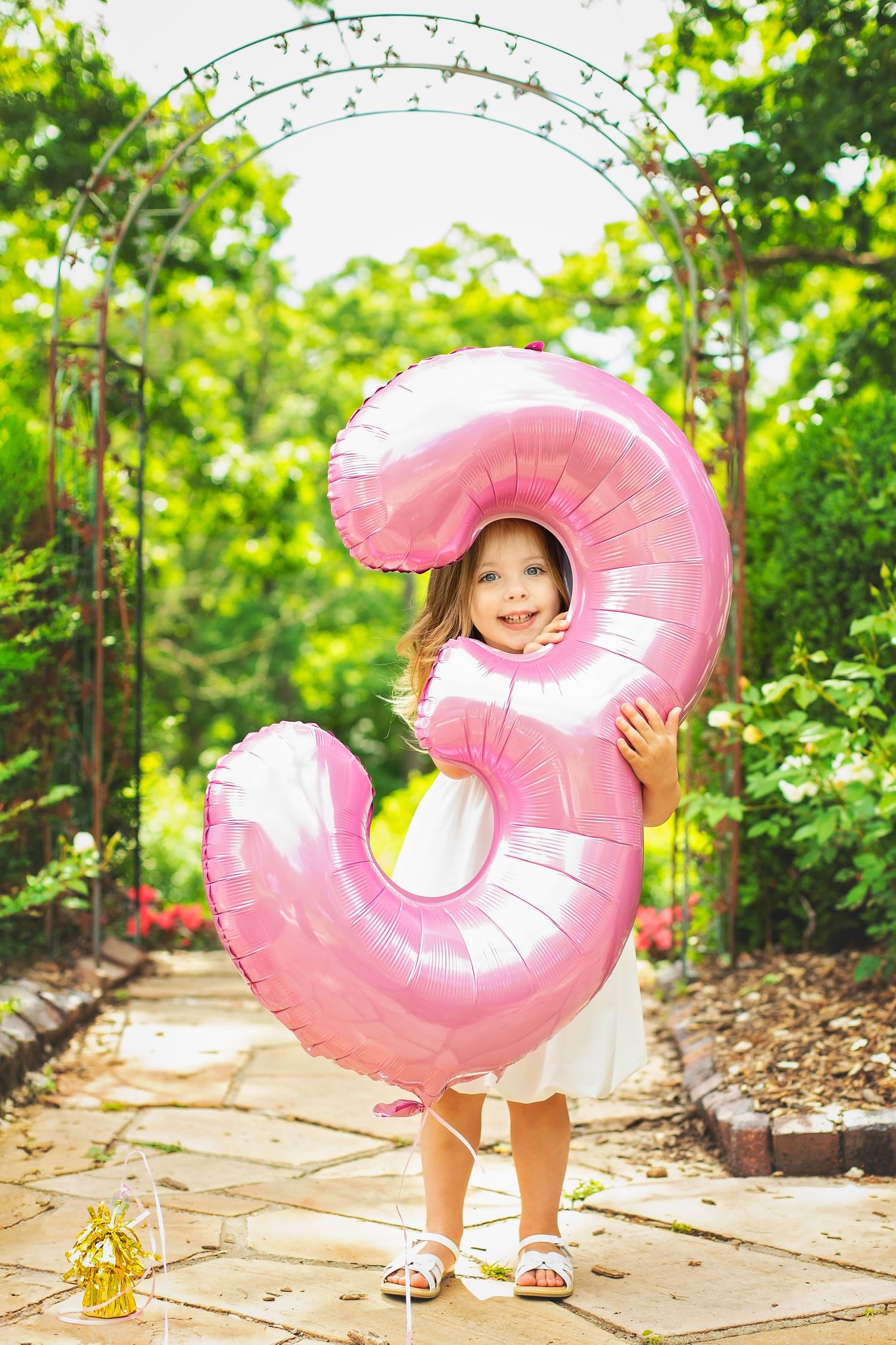 Joyful little girl in white dress holding a large pink number 3 balloon in a lush garden setting.