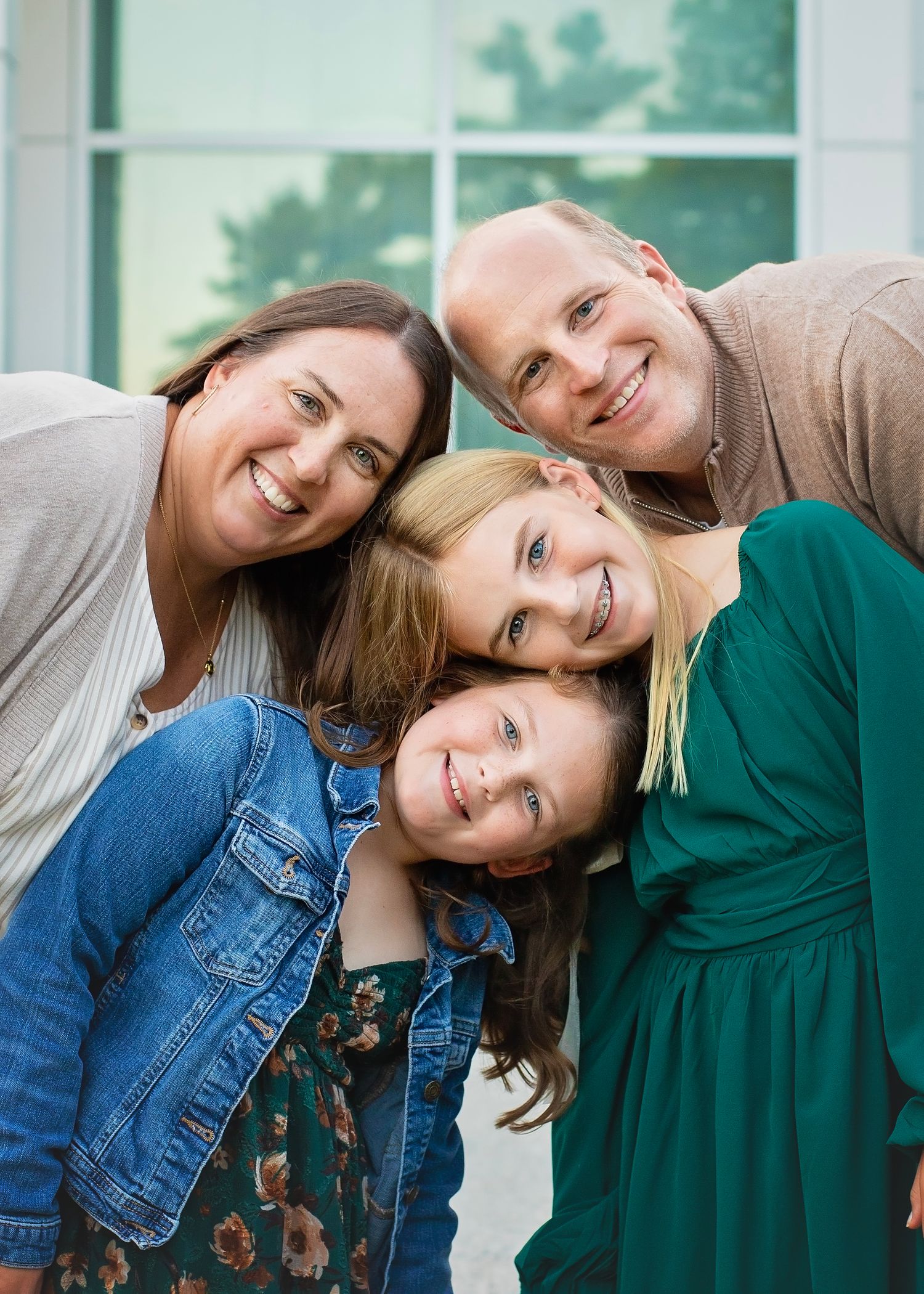 Family of four huddled together cheek to cheek, smiling warmly in a close portrait outdoors.