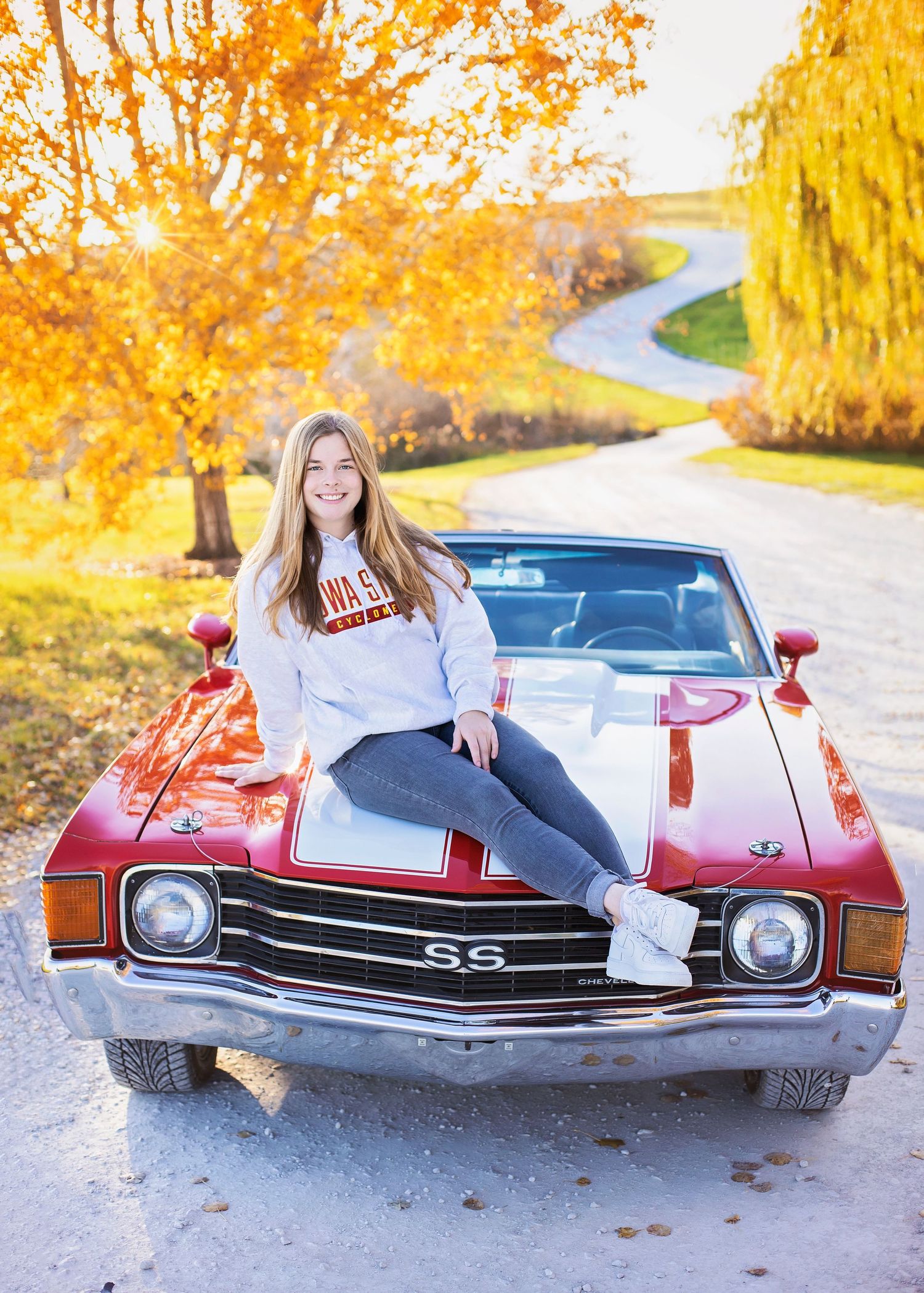 Smiling teen girl sits on red classic Chevy SS car hood surrounded by vibrant autumn foliage.