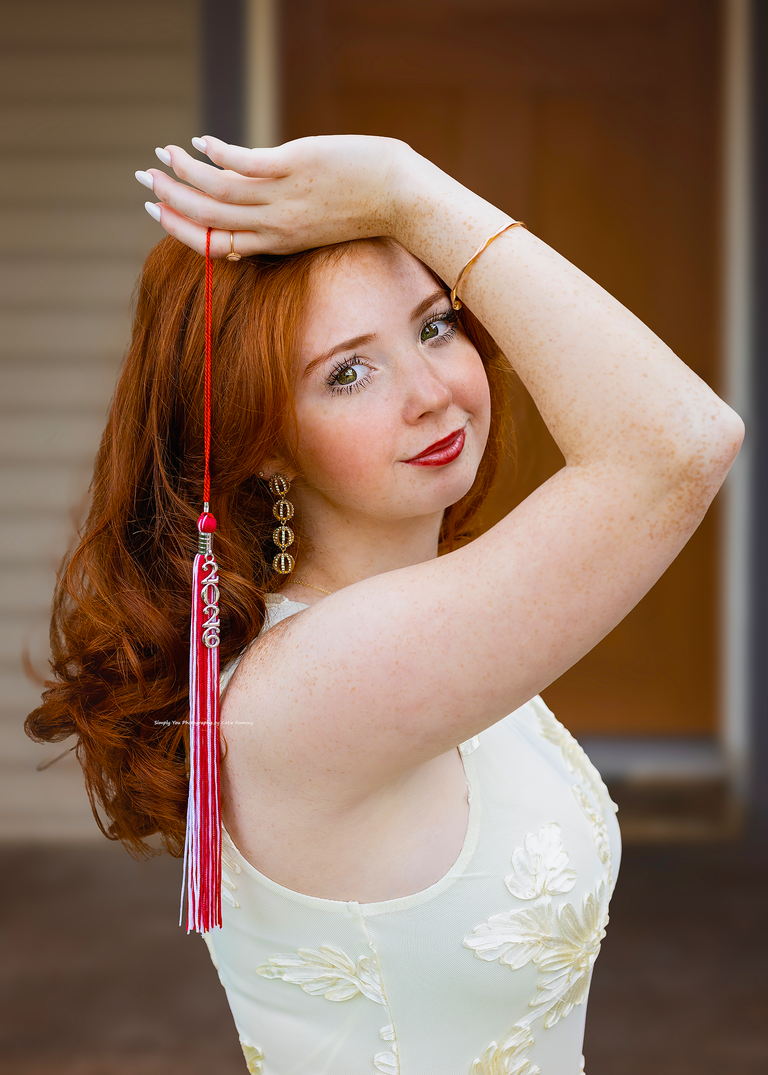 Young redhead woman in white floral dress poses gracefully with arms raised, wearing red tassel earrings outdoors.