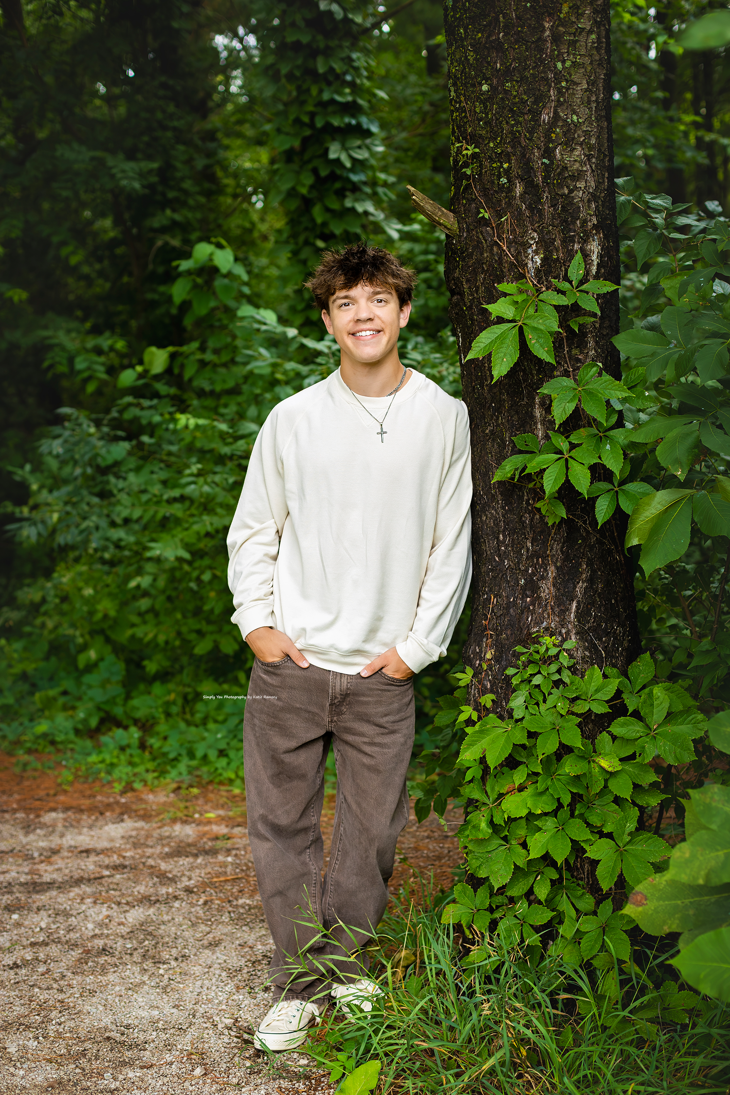 Smiling teen boy in white sweater and gray jeans leaning against a tree in a lush green forest setting.