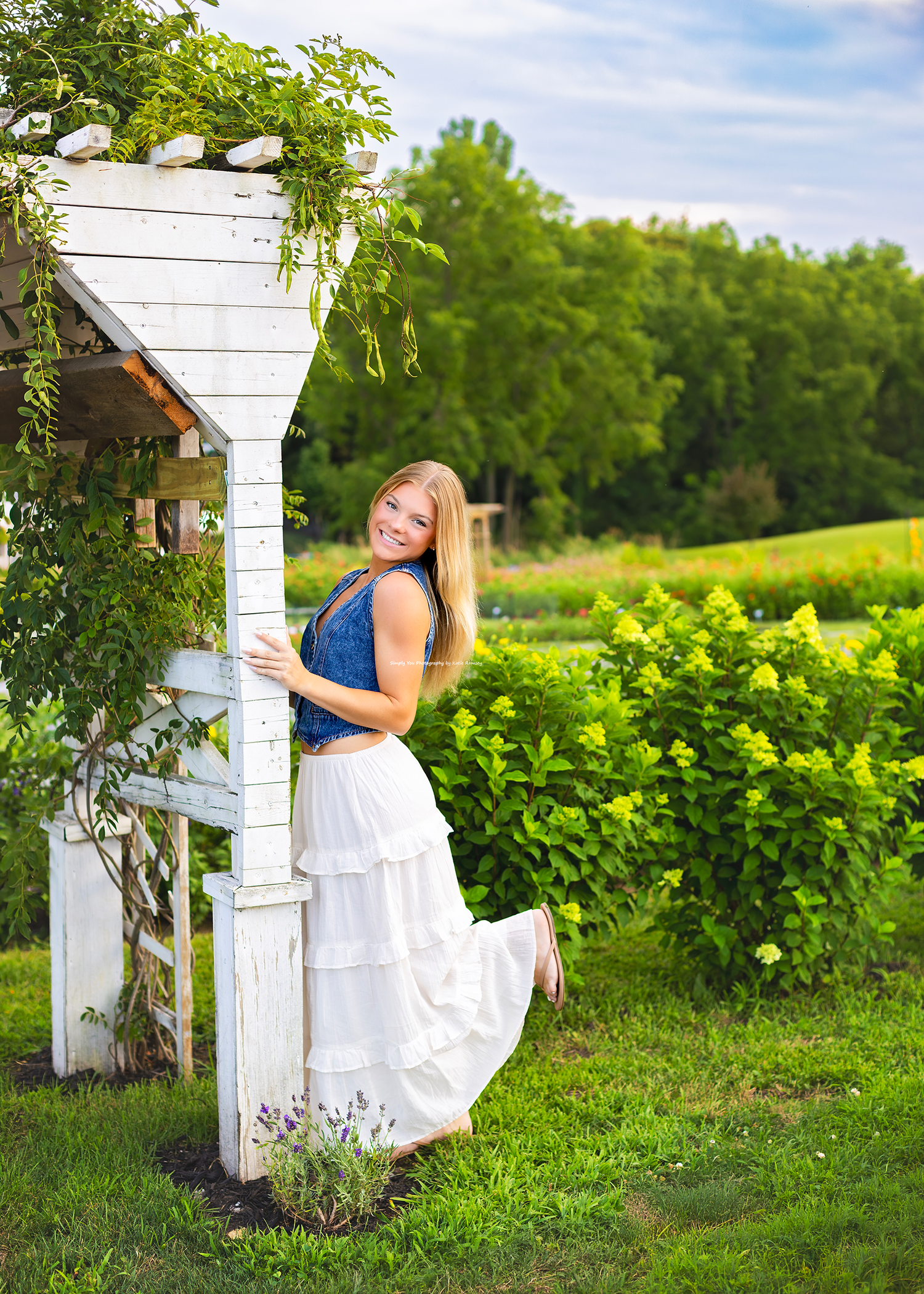 Young woman in white skirt and blue top posing by a white garden arbor surrounded by lush greenery.